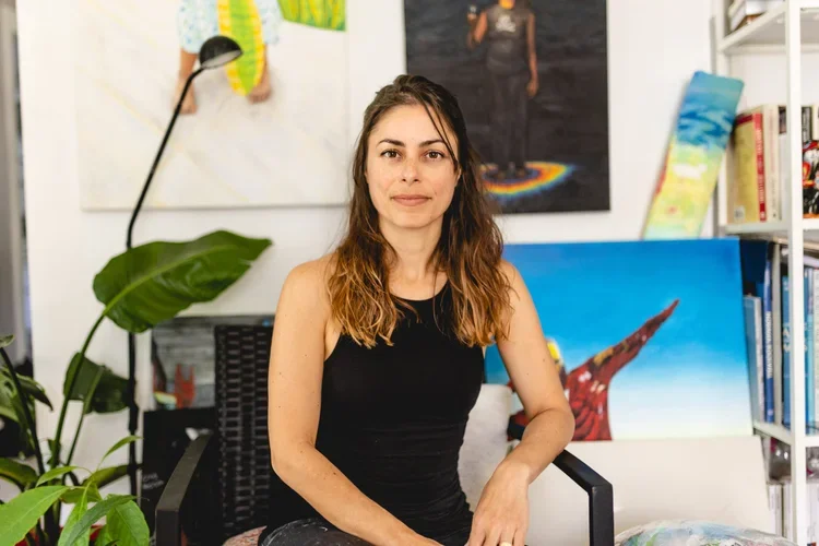 A young woman sitting on a black chair in an art studio or library with paintings and books in the background.