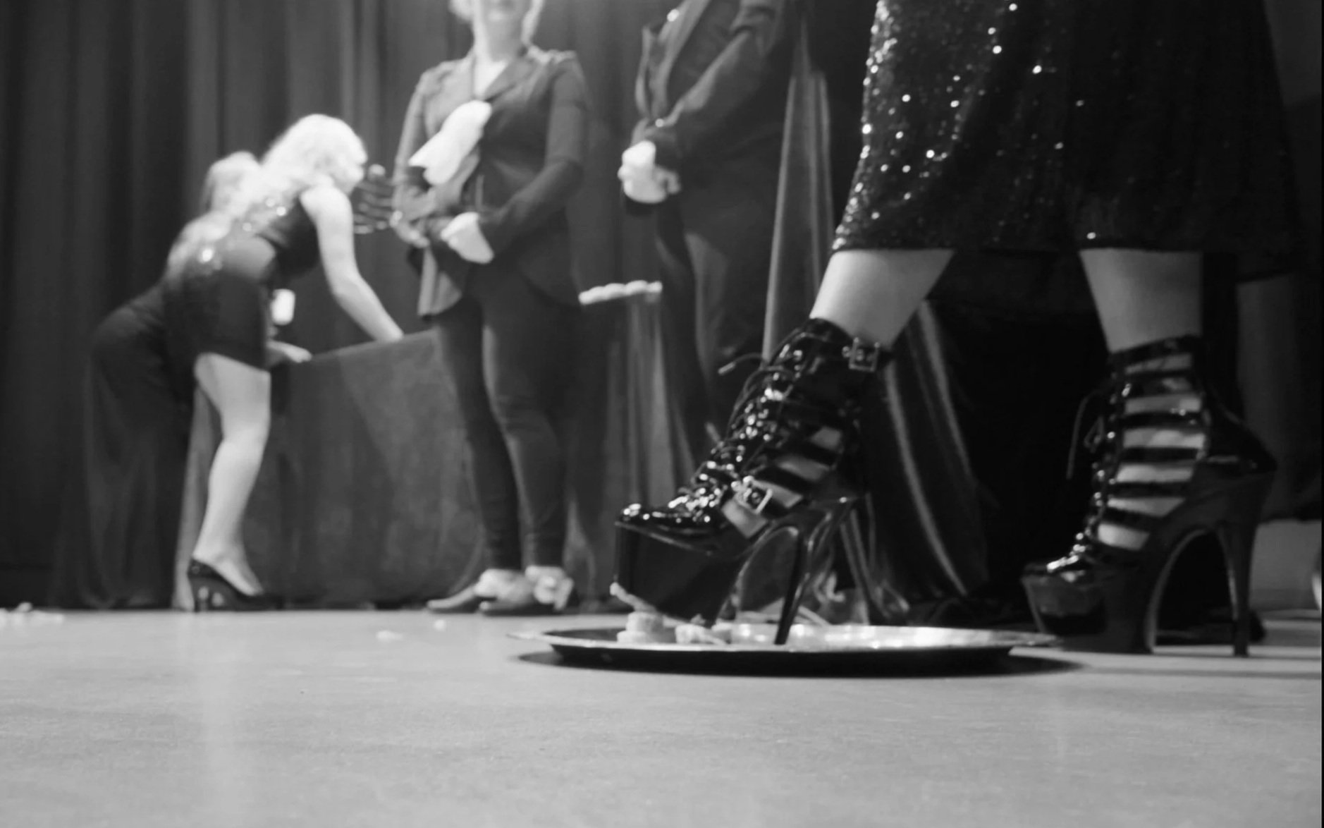 People in costumes on stage, with one person's sequined high-heeled boots and part of their dress prominently in foreground.