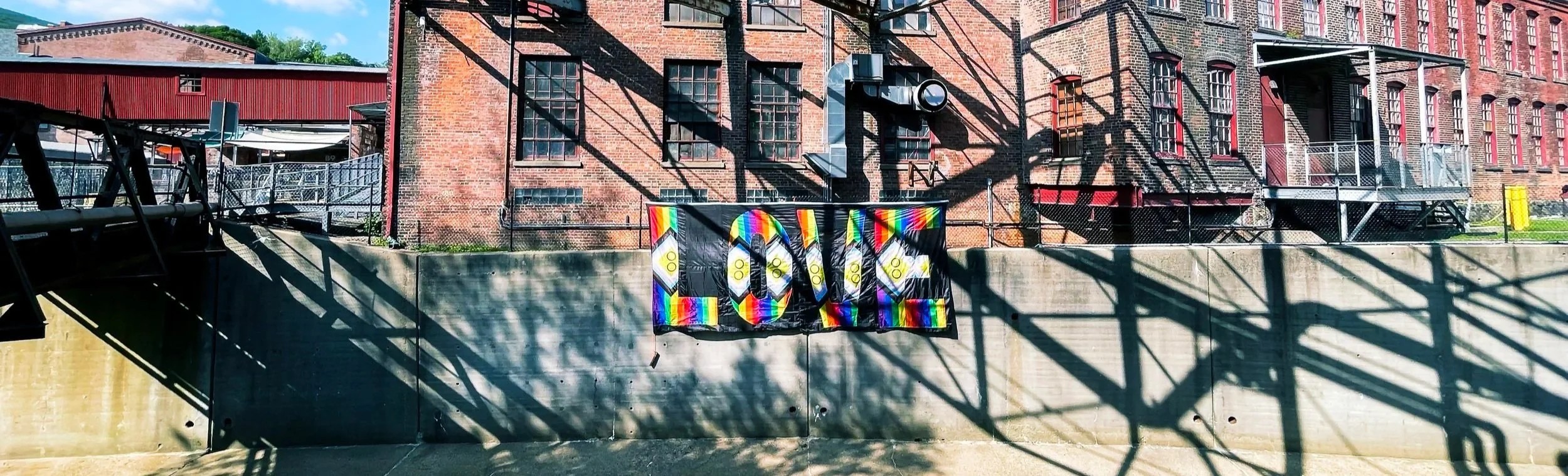 Colorful rainbow flag with a love message hanging on a concrete wall, with shadows of an overhead metal structure and a brick building in the background daytime