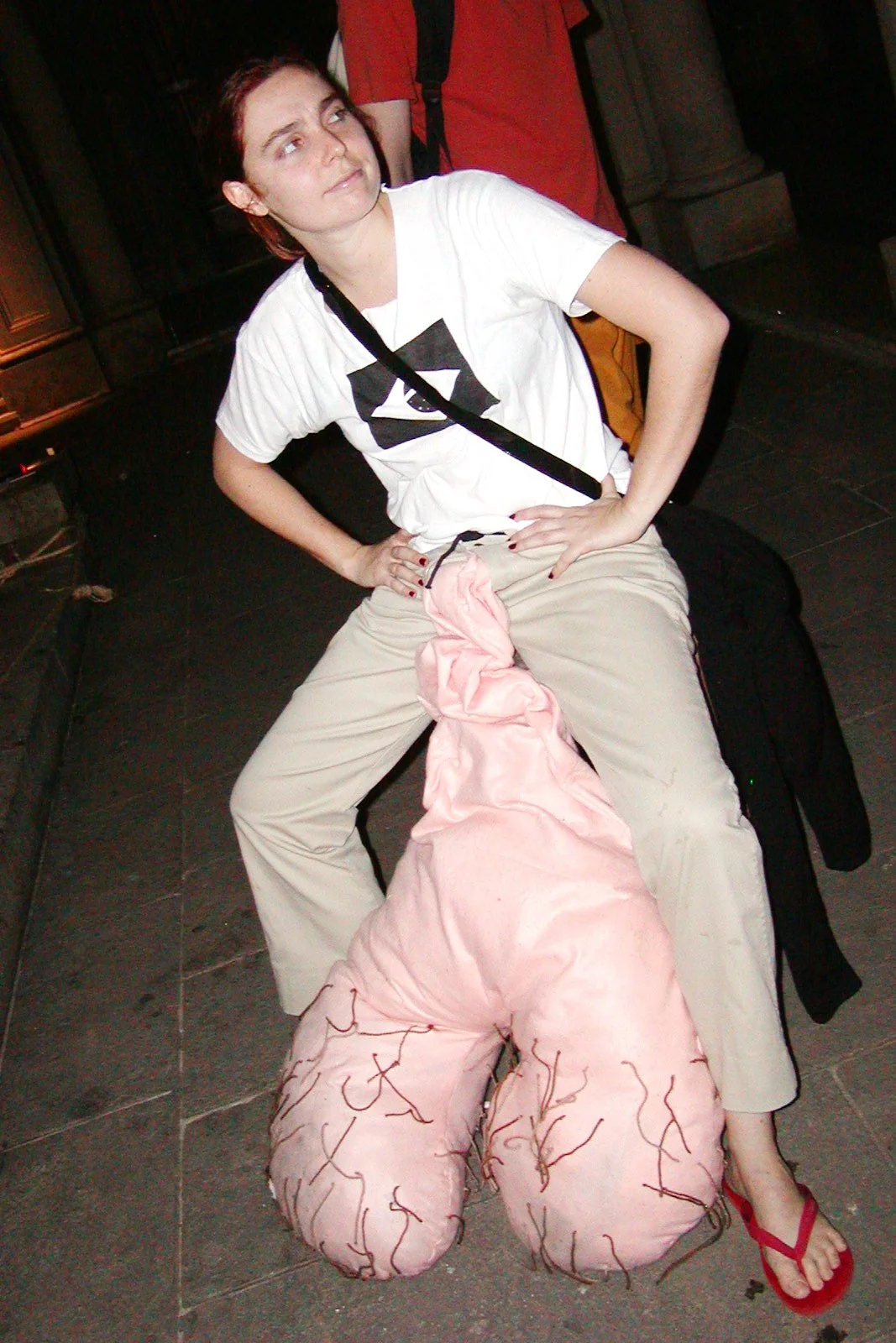 A woman sitting on a black couch with her hands on her hips, wearing a white t-shirt and beige pants, with a pink object resembling a pair of testicles with veins between her legs, in an indoor setting.