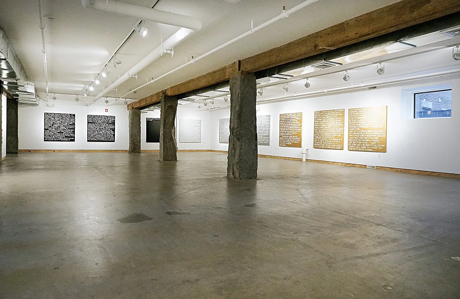 An art gallery with white walls, concrete floors, and exposed wooden and concrete supports. Several pieces of dark and light-colored text-based artwork are displayed on the walls. The gallery is well-lit with track lighting.
