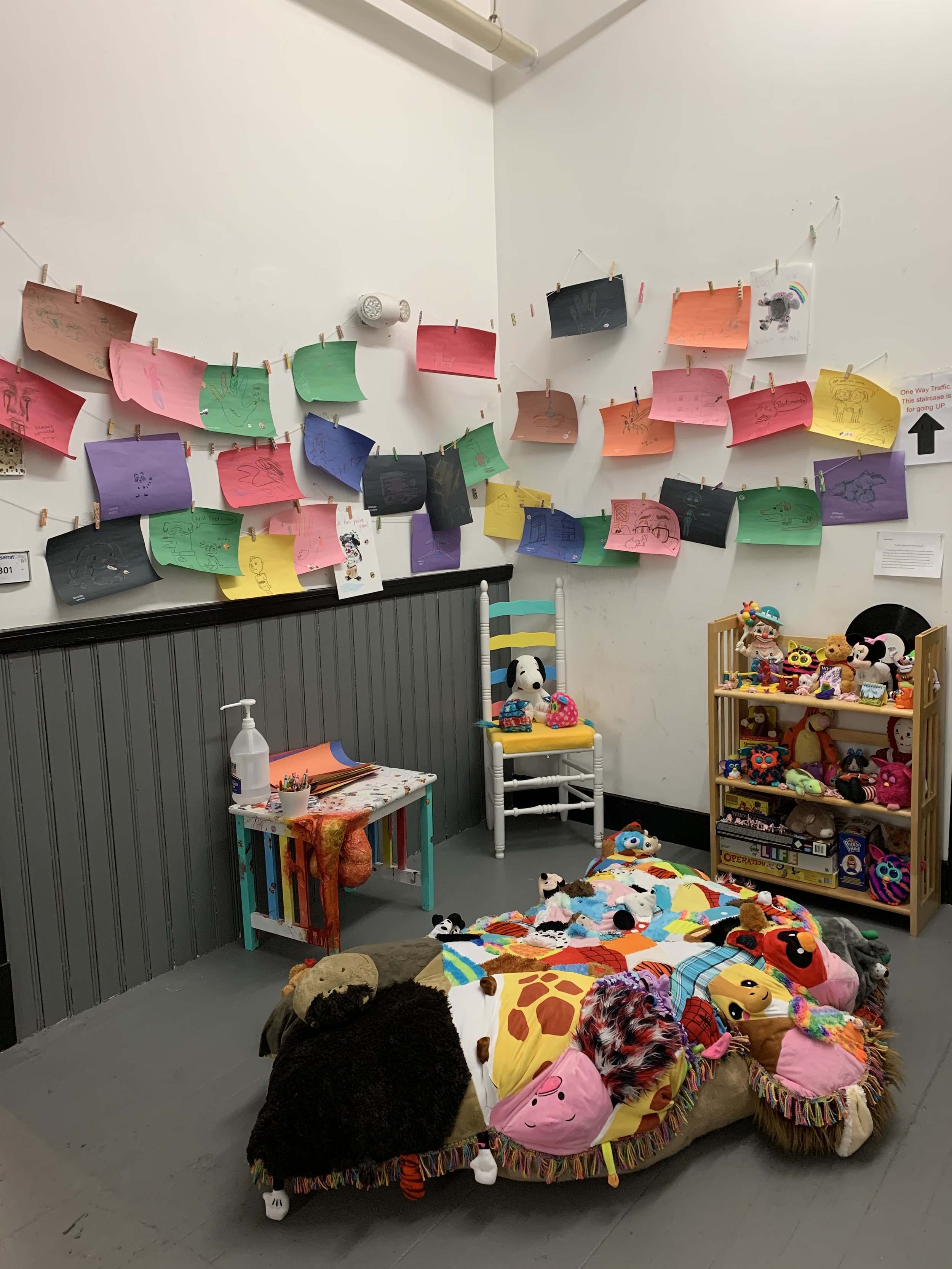 A room decorated with colorful children's drawings on pink, green, purple, yellow, red, orange, and black paper hanging on a string. The room contains a small table with art supplies, a white chair with toy dog, and a wooden shelf filled with plush toys. On the floor, there are various stuffed animals and pillows, resembling a playful and cozy space for children.