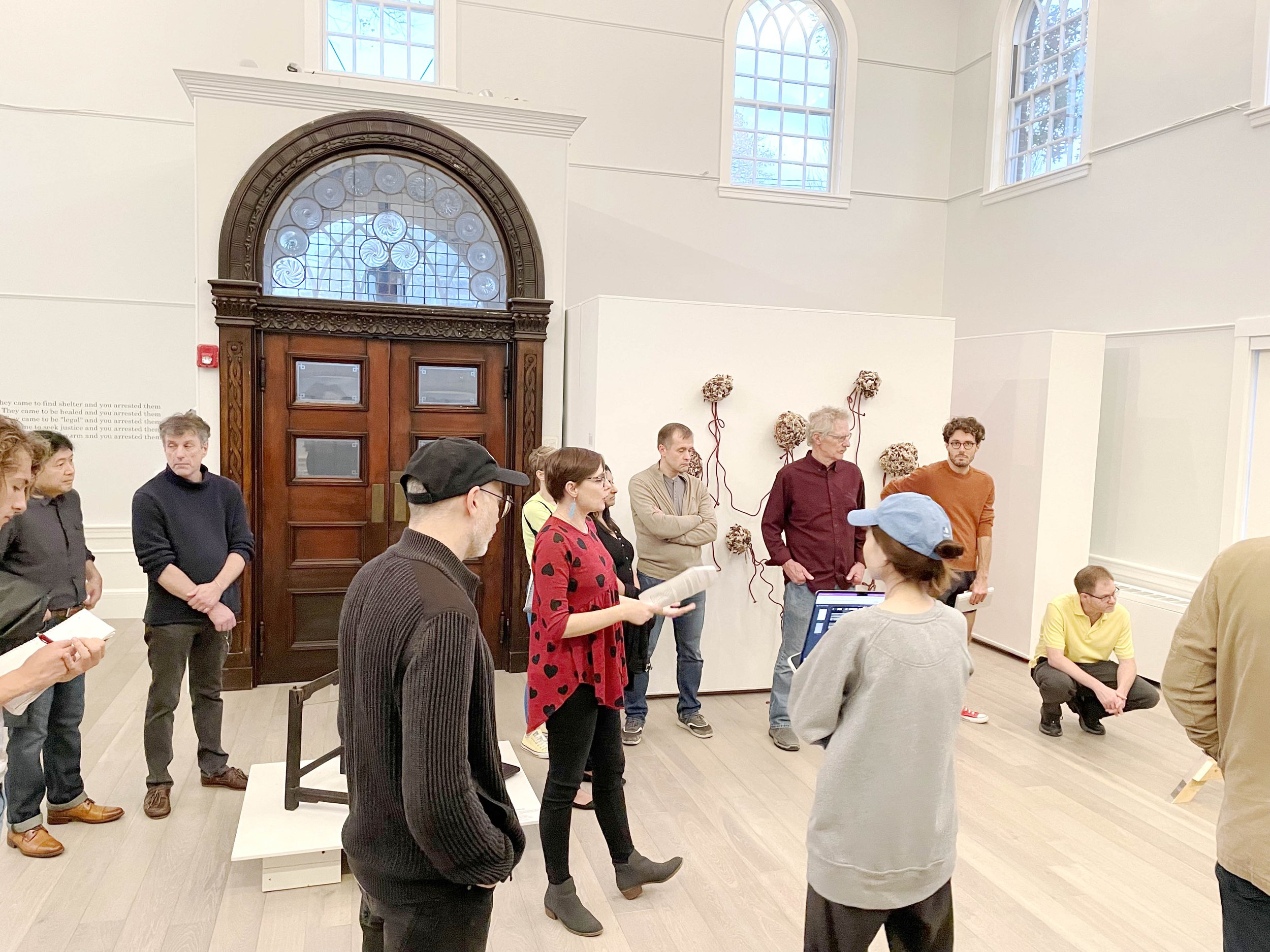 Group of people gathered in an art gallery, listening to a woman with glasses and a red polka dot sweater talk. The gallery has a large wooden door, tall windows, and floral wall decorations.