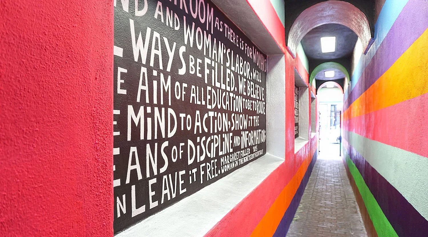 Colorful mural painted on narrow alleyway walls, featuring various bright stripes and a black section with white text.