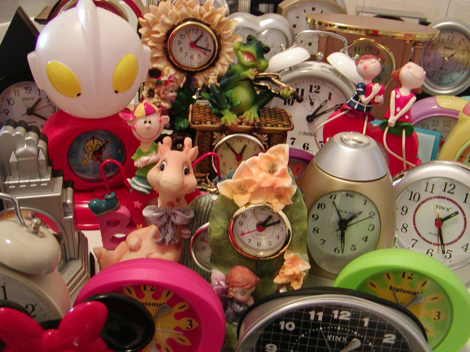 A collection of various colorful clocks and figurines, including animals and cartoon characters, some with decorative elements like flowers and bows, all arranged closely together.