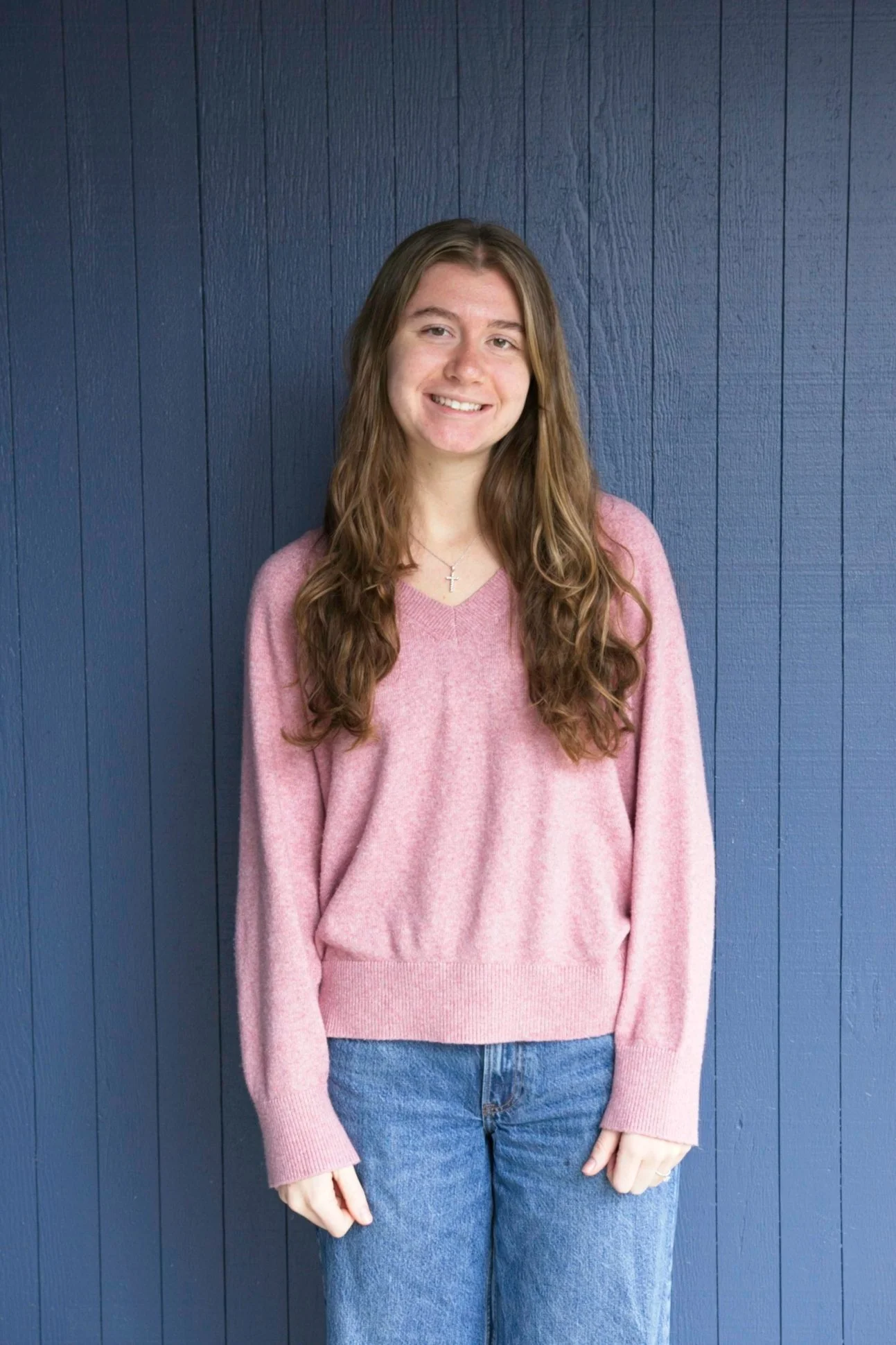 A young woman with long, curly brown hair standing in front of a dark blue wooden wall. She is wearing a pink sweater, blue jeans, and a silver cross necklace, smiling at the camera.