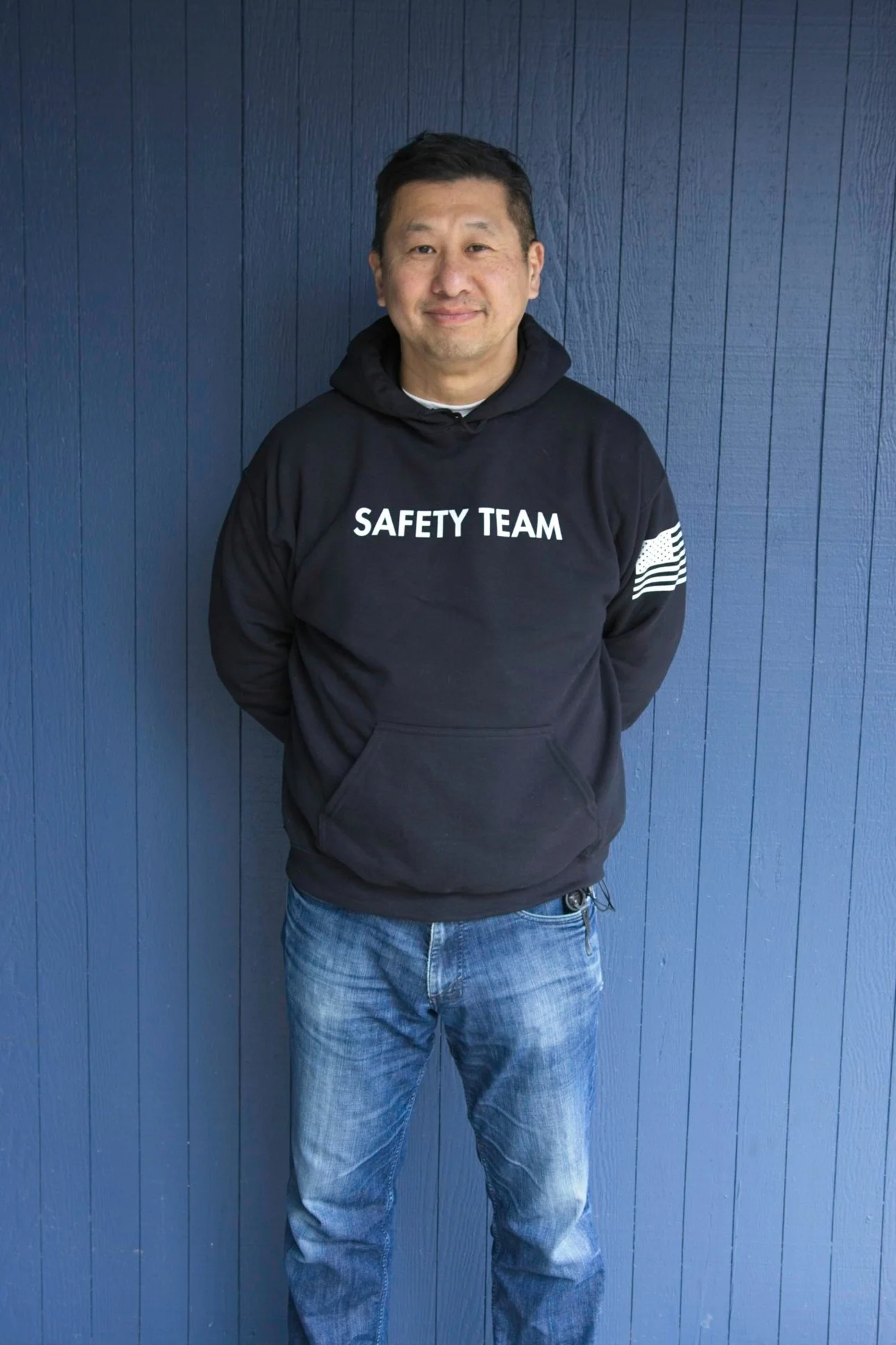 Man wearing a black hoodie with 'SAFETY TEAM' written on it, standing against a blue wooden wall.