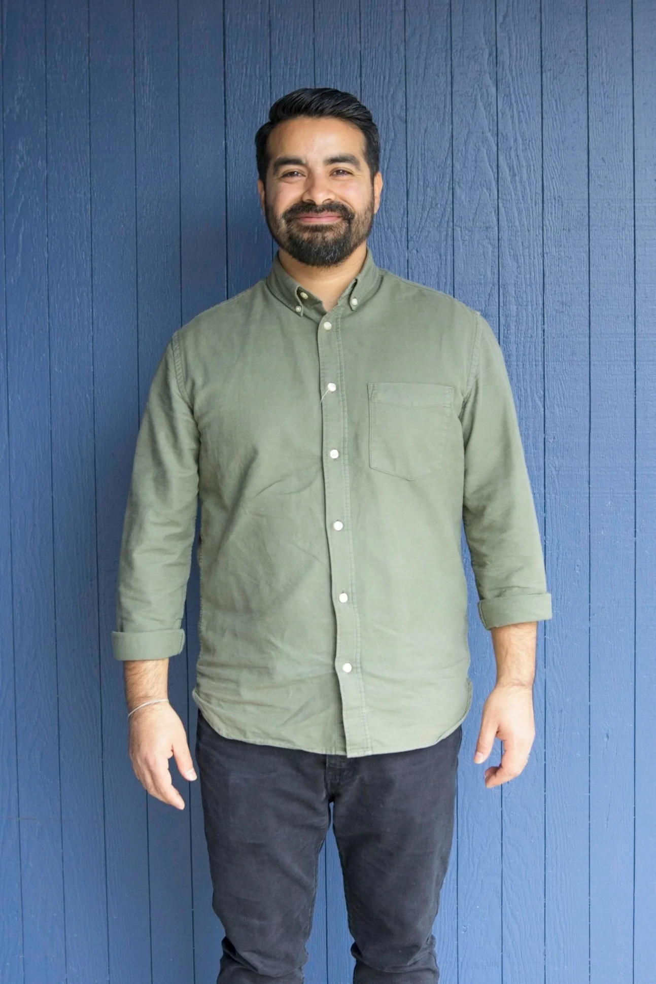 Man with dark hair and beard smiling, wearing a green button-up shirt with rolled sleeves, standing against a blue wooden wall.