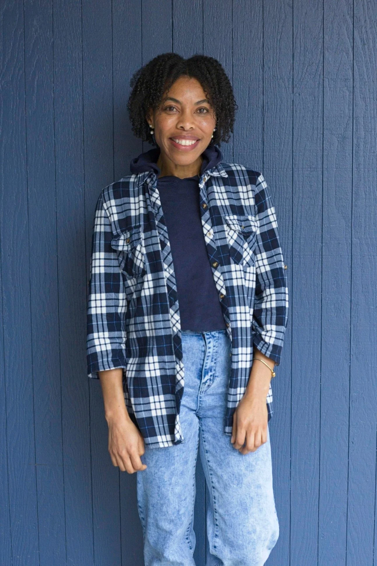 A woman with short curly hair smiling and standing against a dark blue wooden wall. She is wearing a navy hoodie, a blue and white plaid flannel shirt, and light blue jeans.