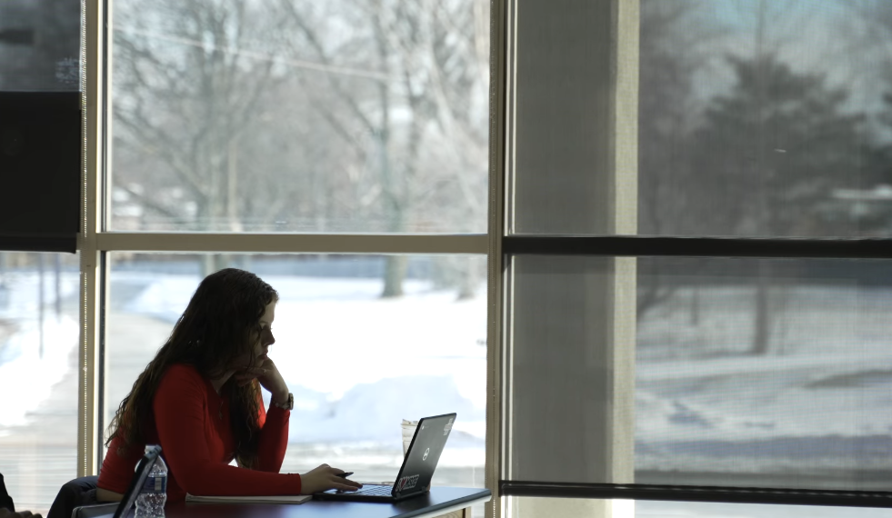 A student works on a laptop by broad windows reflecting a snowy landscape