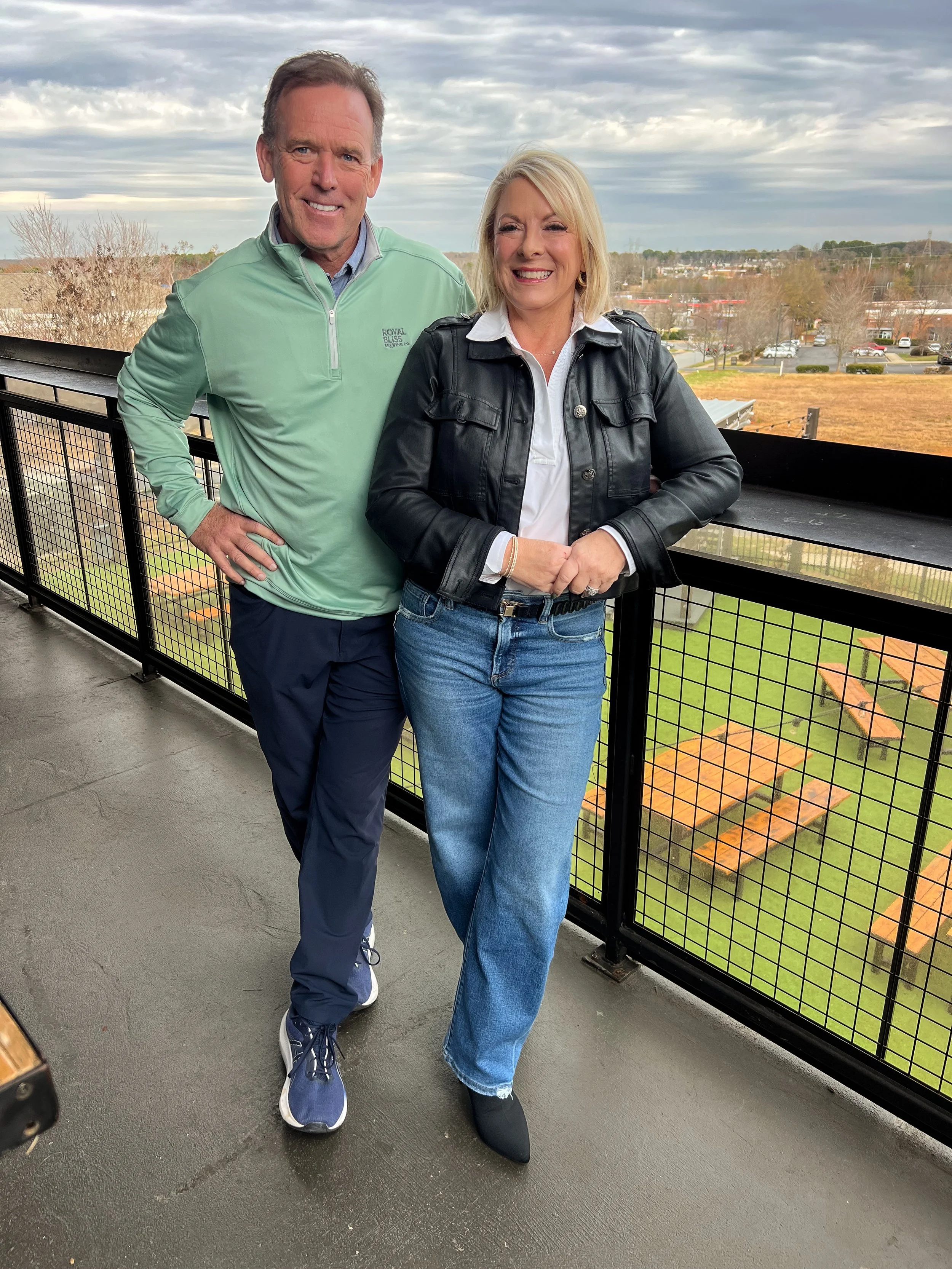 Founders and Owners of Royal Bliss Brewing, Larry and Ginger Griffin standing on the terrace. The man is wearing a light green jacket, navy pants, and sneakers. The woman is wearing a black leather jacket, white shirt, blue jeans, and black shoes.