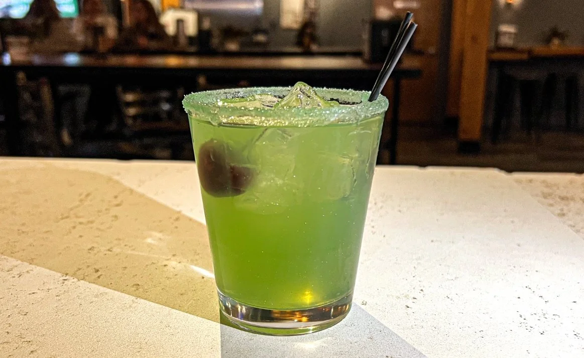 The Grinch Who Stole Blissmas cocktail from Royal Bliss with tequila, melon liqueur, simple syrup and lime juice with edible glitter and a cherry garnish.