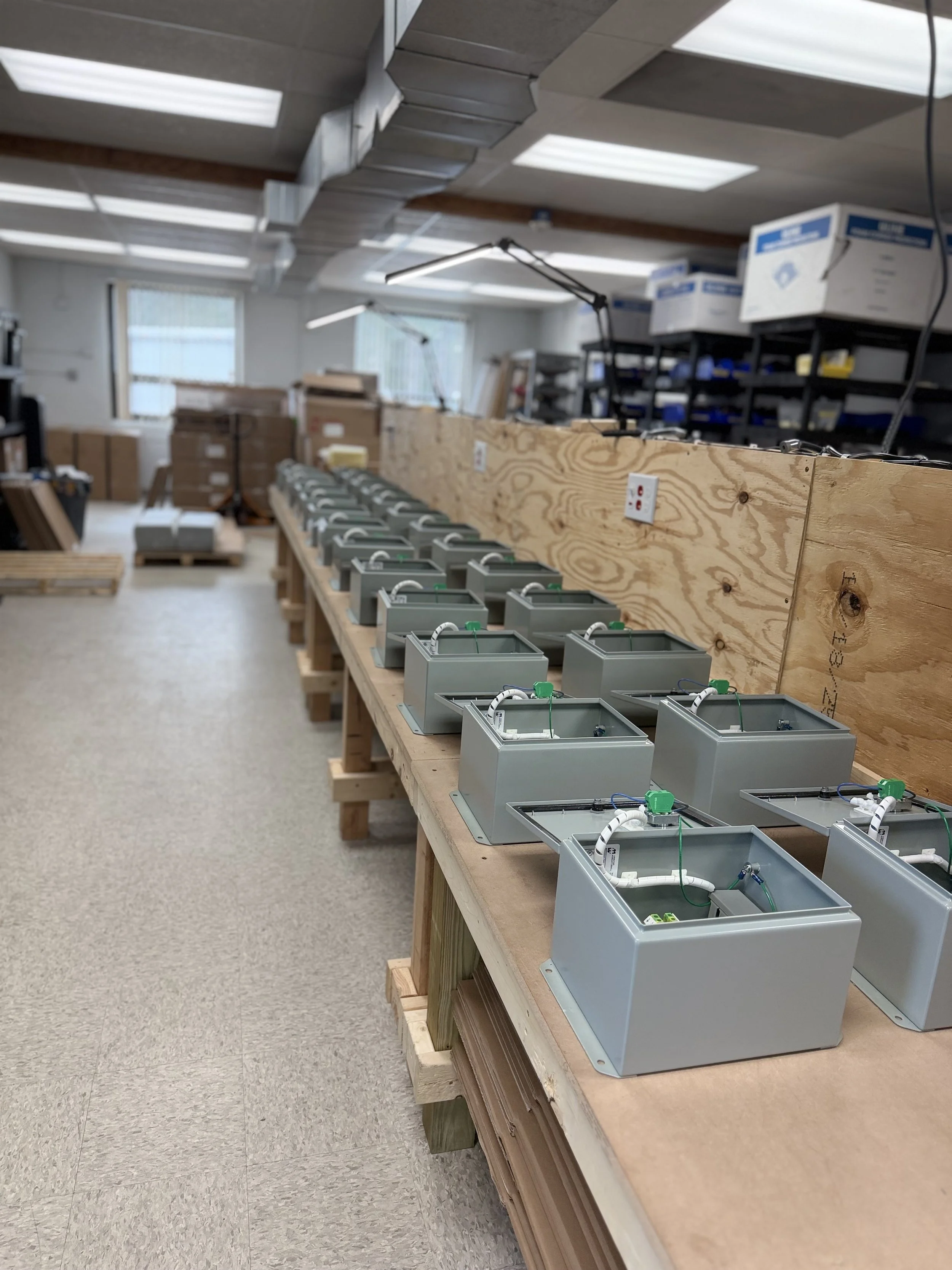 An industrial workspace with multiple electrical boxes and wiring on a long wooden workbench, with shelves and boxes in the background and overhead fluorescent lighting.