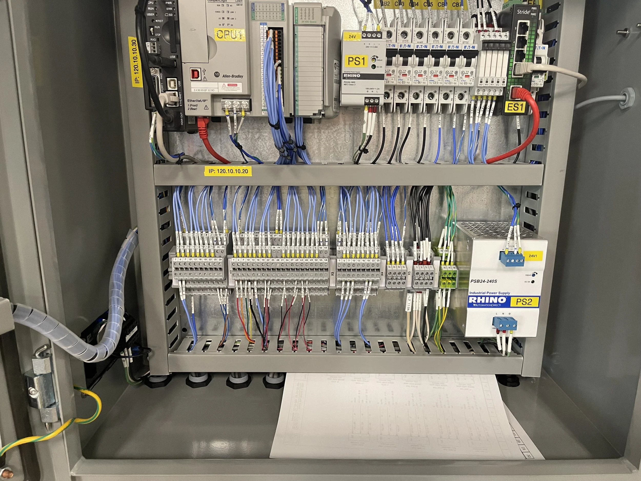 An electrical control panel with various wires, circuit breakers, and components labeled with yellow tags, including CPU, PS1, ES1, and IP address, with a printed sheet of technical diagrams at the bottom.