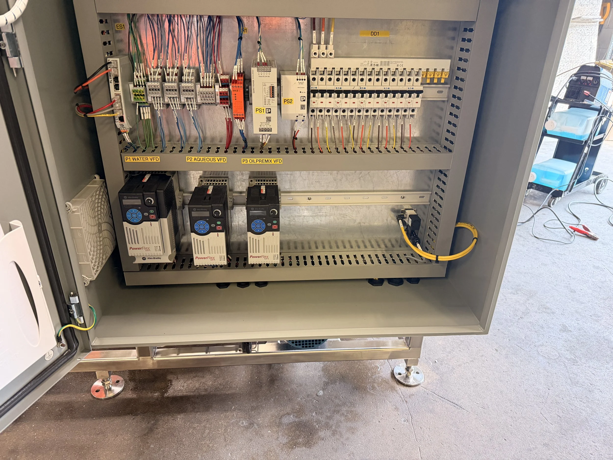 An electrical control panel with various wires, circuit breakers, and electrical components inside a metal cabinet. The cabinet is labeled with specific labels such as P1 WATER VFD, P2 AQUEOUS VFD, and P3 OILPREMIX VFD.
