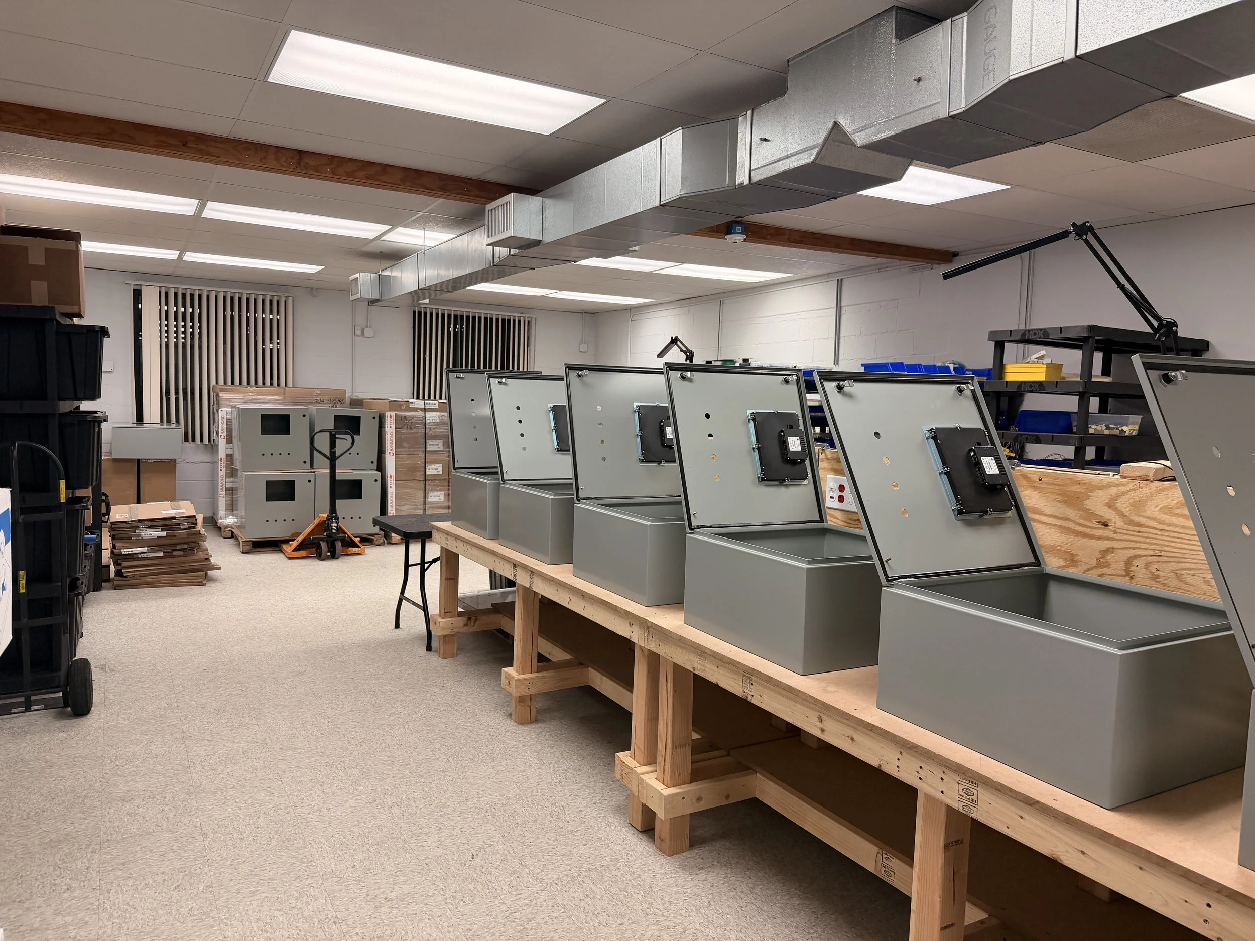 Workshop or manufacturing space with metal enclosures, workbenches, boxes, and equipment.