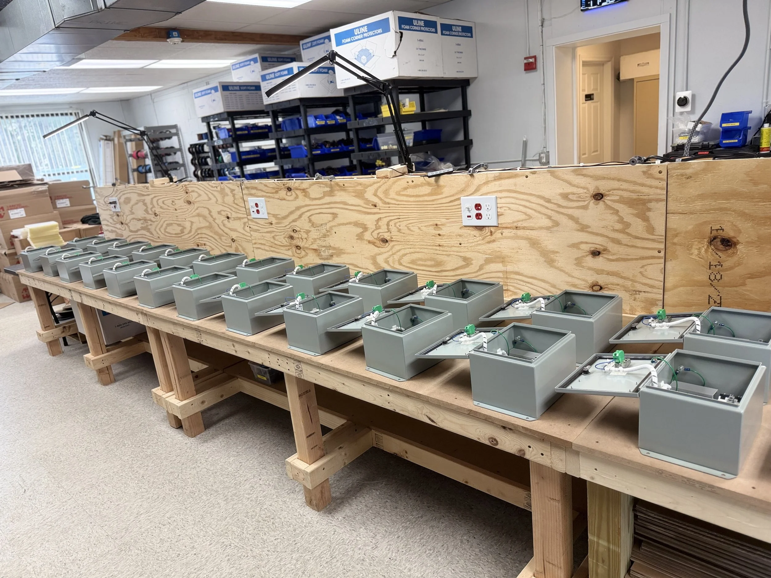 An indoor workspace featuring a long wooden table with multiple metal boxes connected to electronic components. In the background, there are shelves filled with blue storage bins, boxes labeled 'ULINE FOAM Corner Protectors,' and a wall with electric