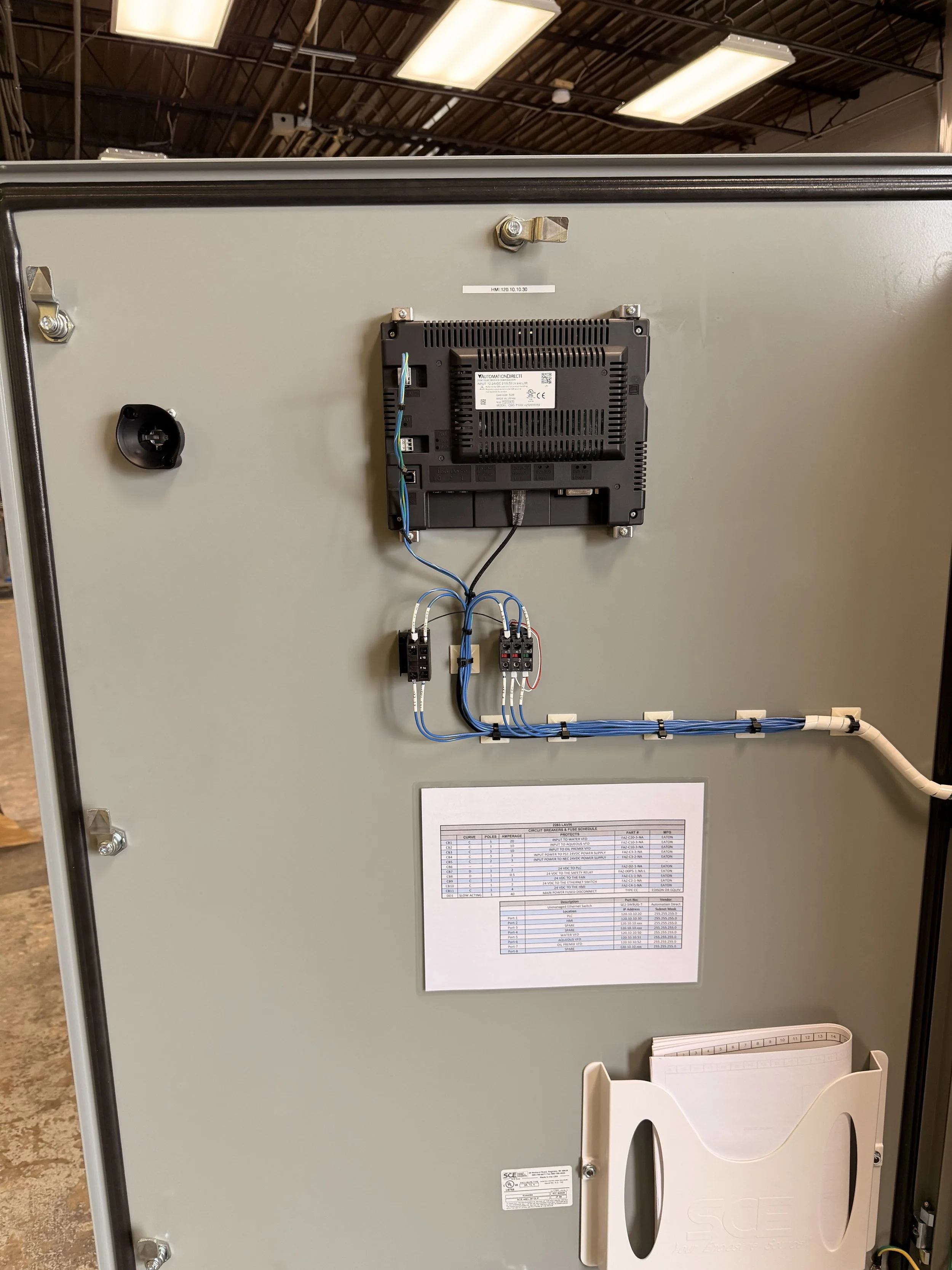 Back side of an electrical control panel with wiring, a display, and a paper schedule attached, set in an industrial environment.