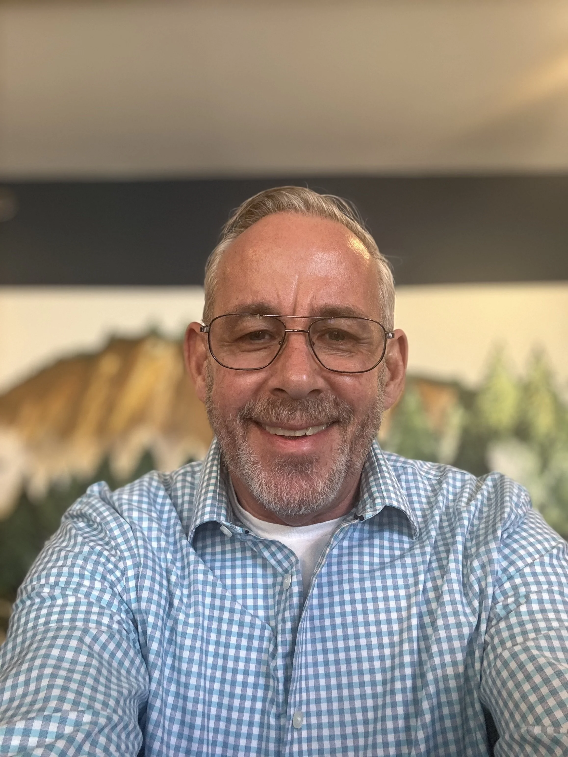 A smiling middle-aged man with glasses and a beard taking a selfie in an indoor setting with artwork and plants in the background.