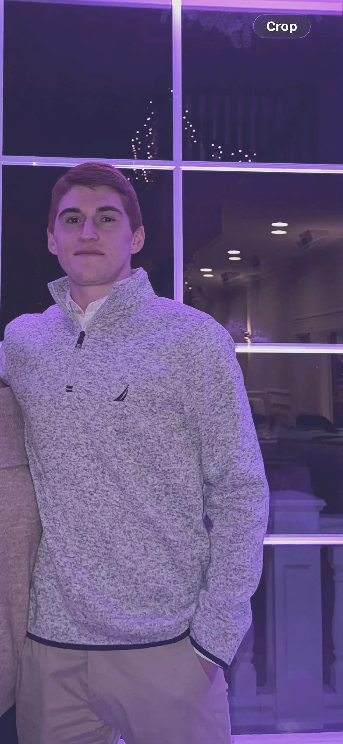 A young man in a light patterned quarter-zip sweater and beige pants stands indoors at night, with ceiling lights and a window with reflections behind him.