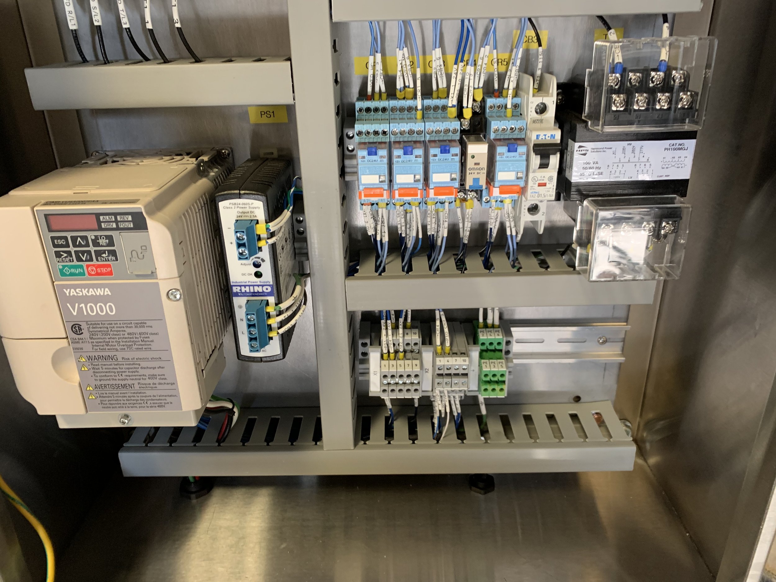 An electrical control panel with various components including a Yaskawa variable frequency drive, a power supply, relay modules, terminal blocks, and wiring organized inside a metal enclosure.