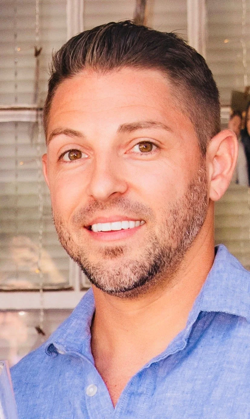A man smiling standing next to an open electrical control panel with wires and components inside, in a workshop with an American flag on the wall.