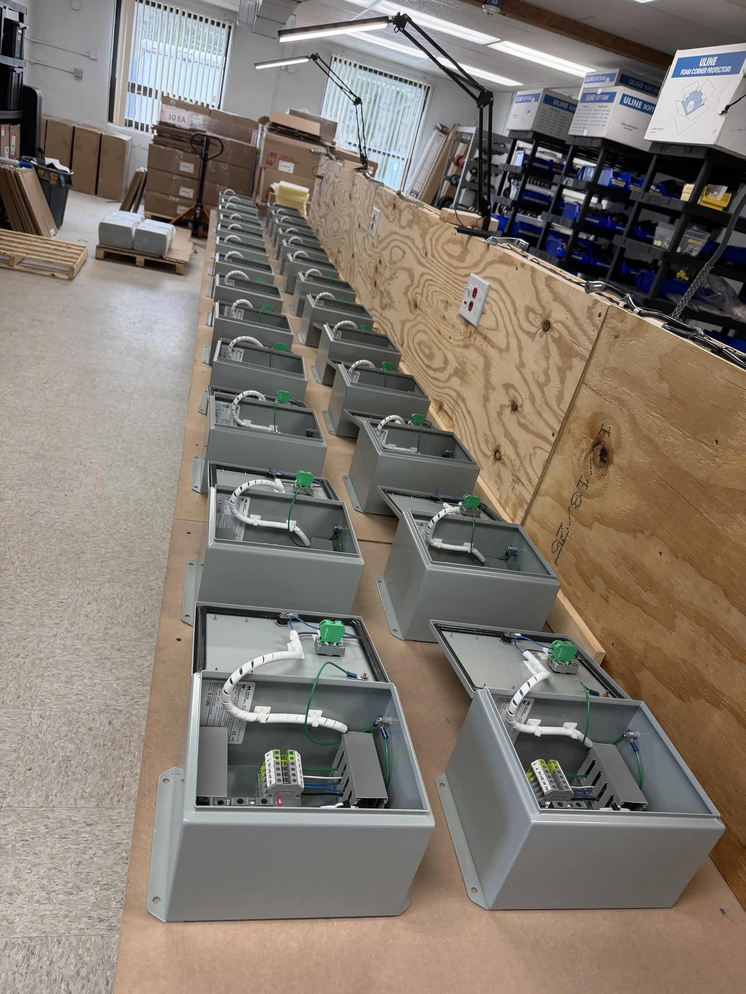 Open electrical control boxes with wiring inside, lined up on a workbench in a workshop, with tools and equipment in the background.