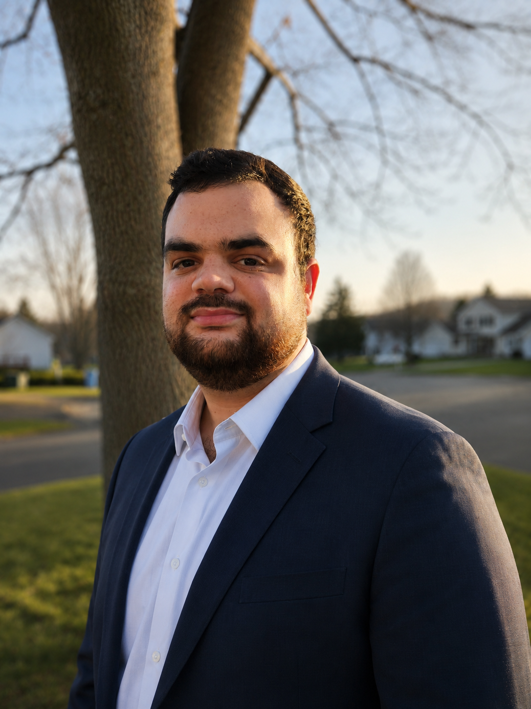 A man in a navy suit and white shirt standing outdoors in front of a tree, with a suburban neighborhood in the background during sunset.