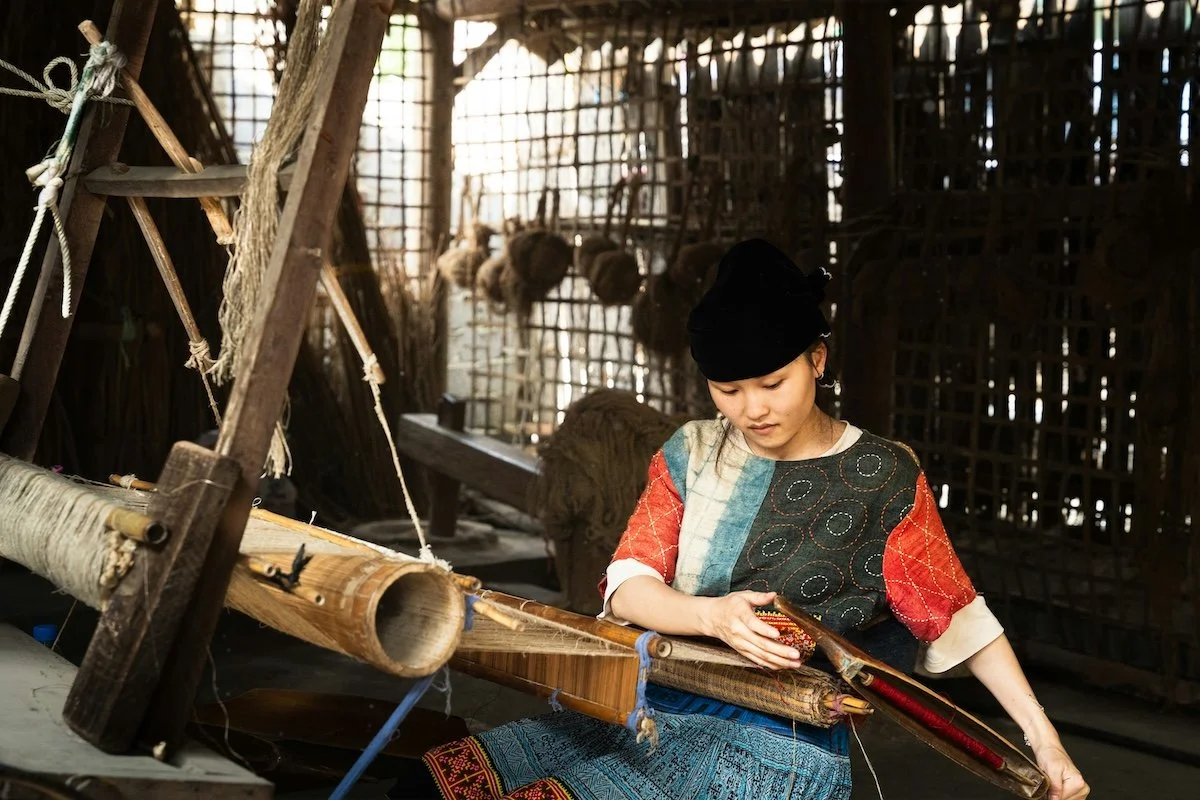 An artisan in traditional clothing operates a large, complex wooden loom, meticulously weaving a textured textile from raw, natural hemp fibers, demonstrating the value of slow and authentic craftsmanship.