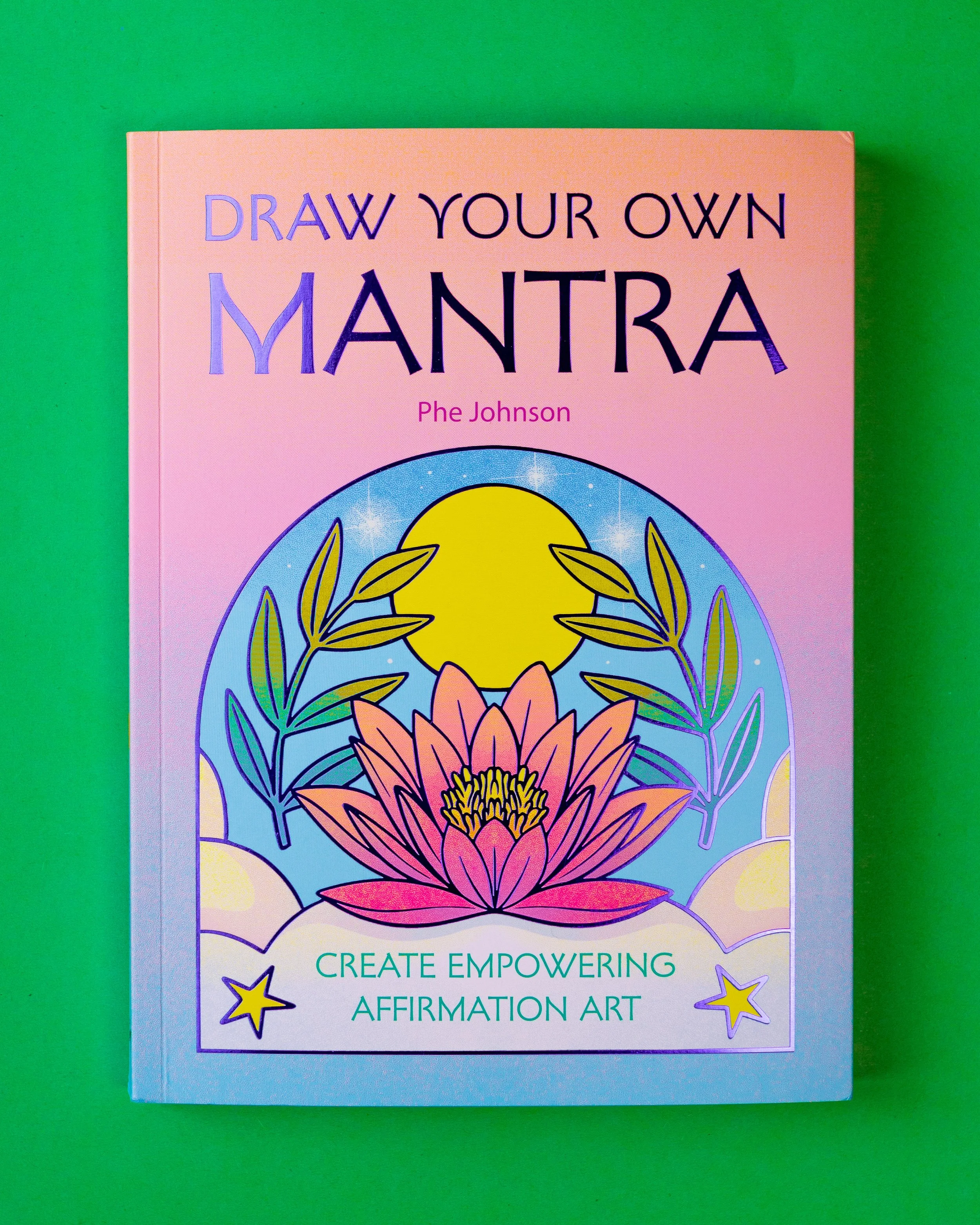 Book titled "Draw Your Own Mantra" by Phe Johnson with colorful cover featuring a lotus flower, sun, and stars on a pink and purple background.