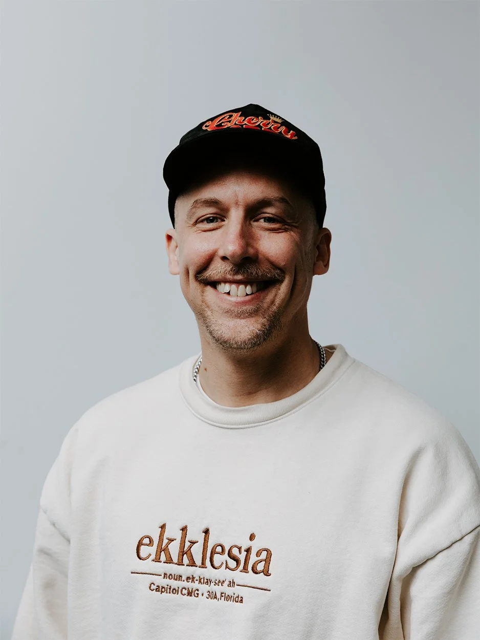 Smiling man wearing a black cap with 'Madhappy' written on it, a white t-shirt, a black jacket with a beige zipper, and a silver chain necklace, standing in front of a brick wall.