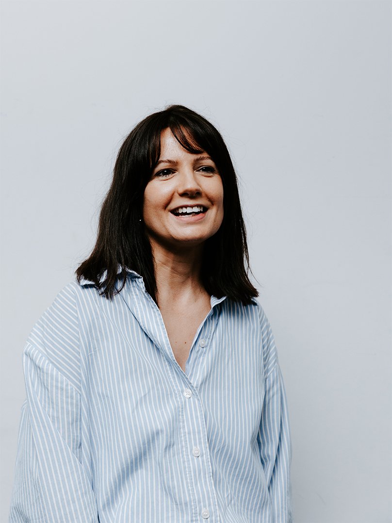 A woman with shoulder-length dark hair wearing a dark denim shirt standing against a plain white background.