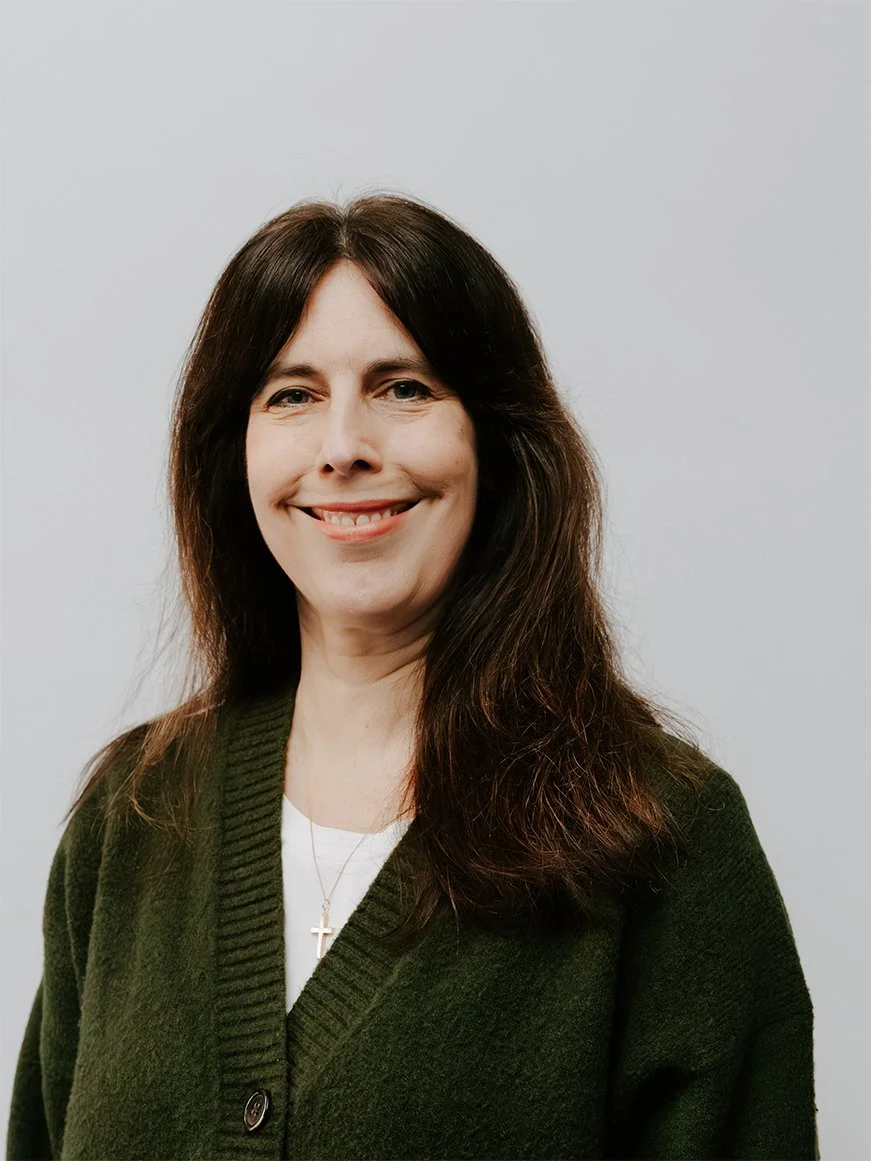 A woman with long dark hair wearing a white shirt smiling and looking slightly upwards, against a plain light-colored background.