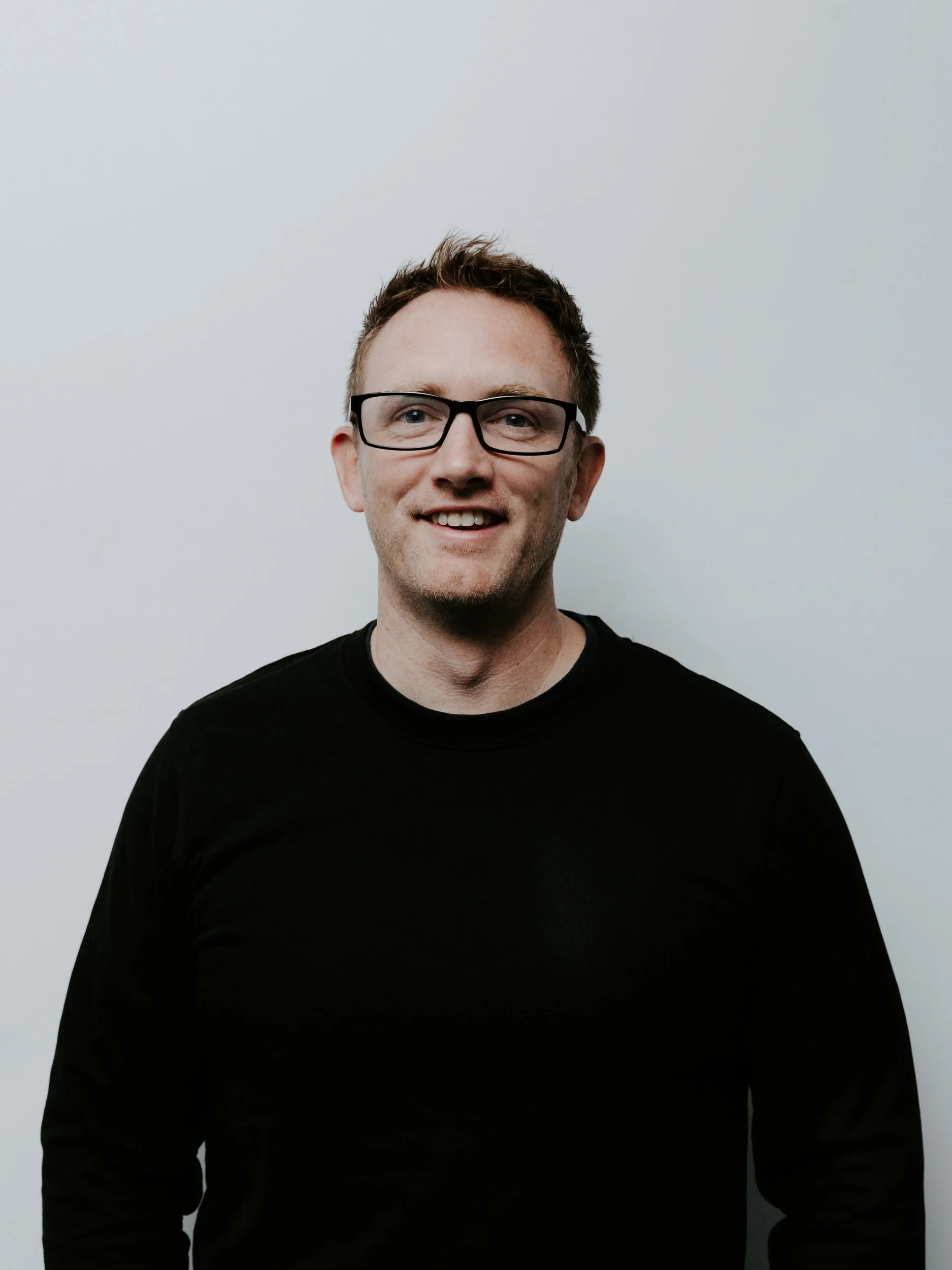 A smiling man with glasses and short, curly hair, wearing a gray sweatshirt, standing against a plain white wall.