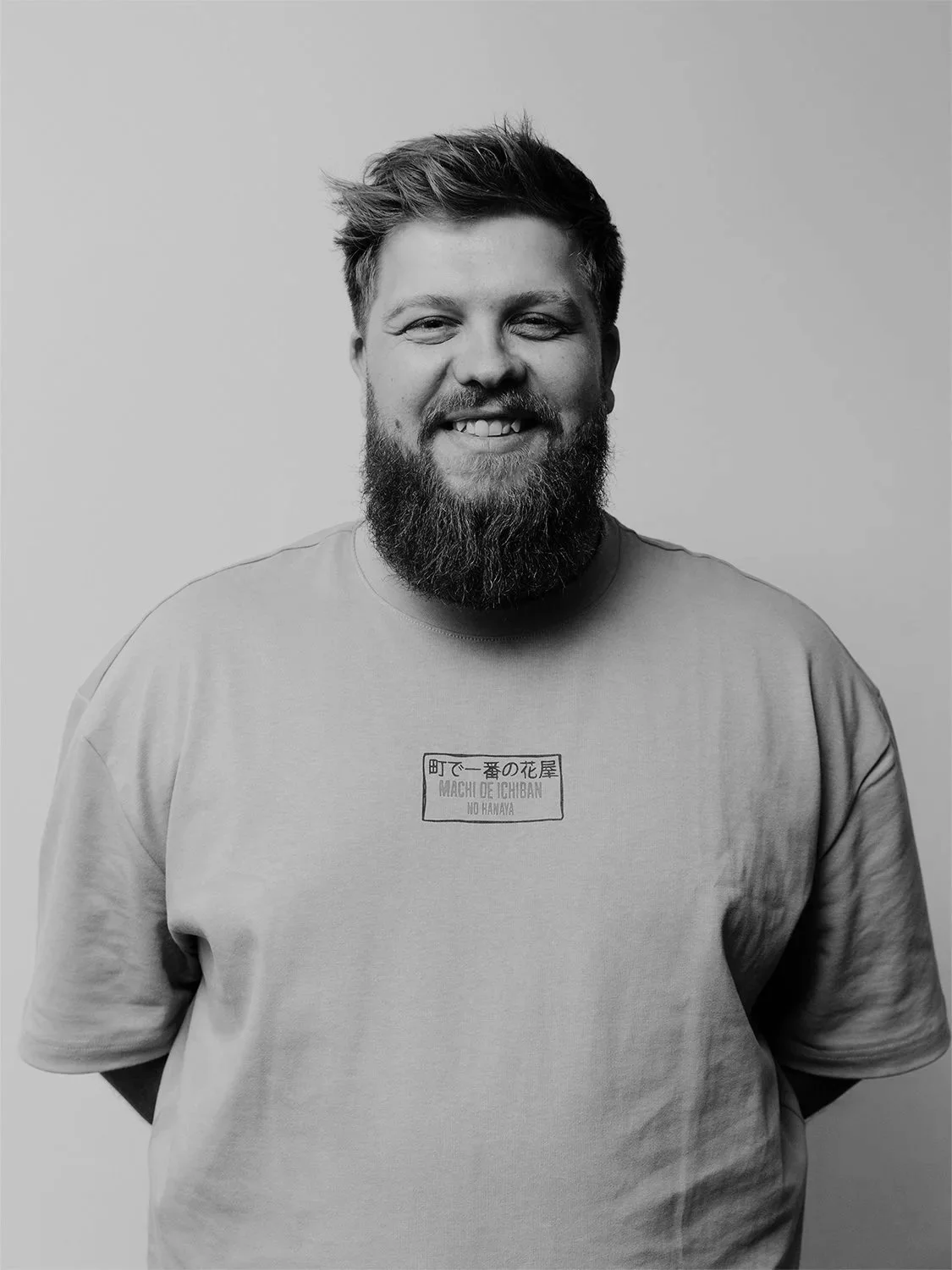 A man with dark hair and a beard smiling while standing in front of a brick wall.