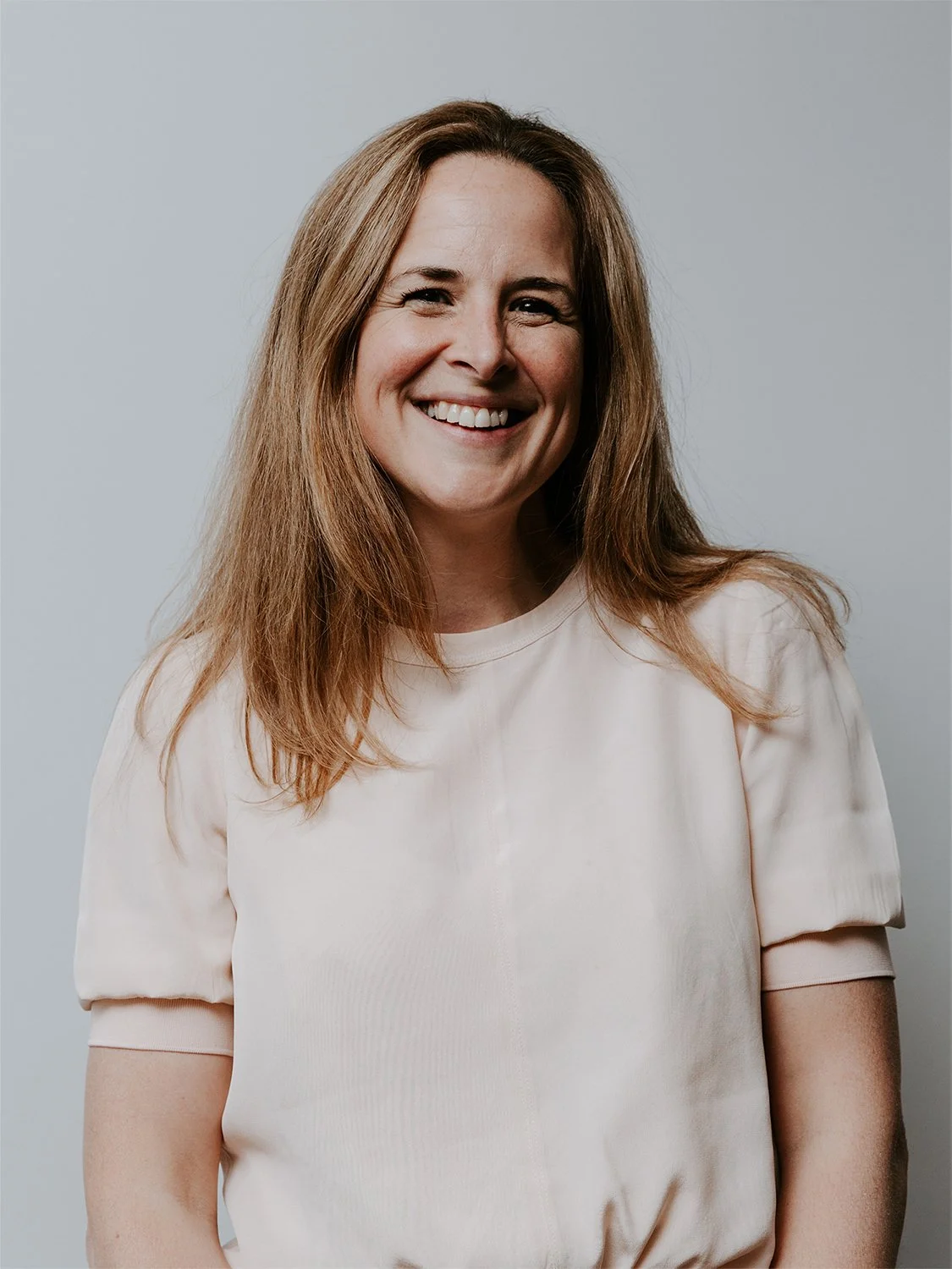 A woman with long, wavy brown hair smiling in front of a red brick wall, wearing a black sweater with white star patterns.