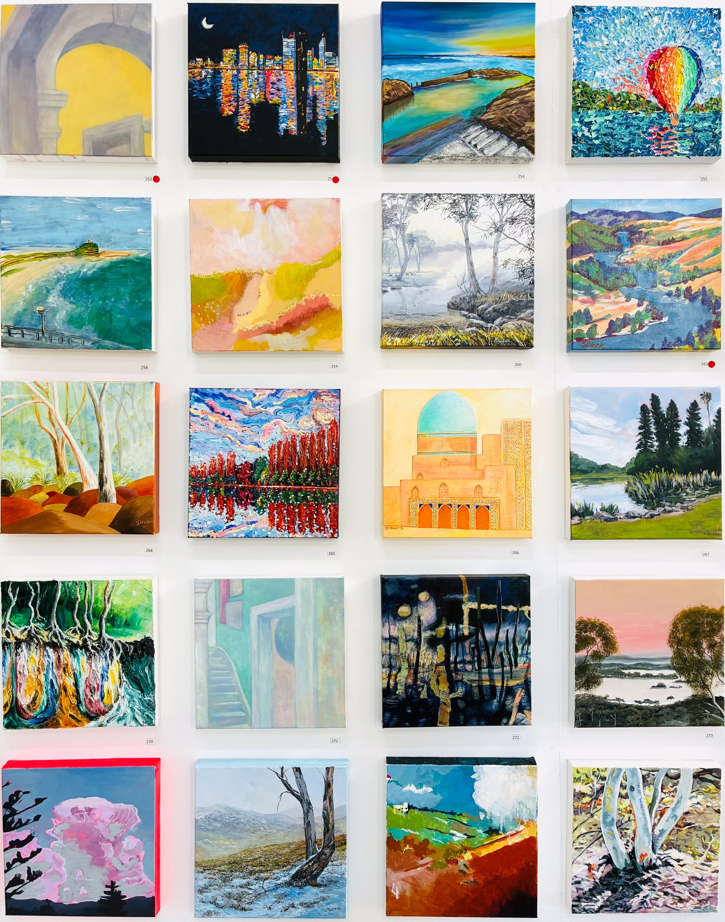 A grid of 20 square paintings hanging on a wall. Mostly colourful landscapes. 