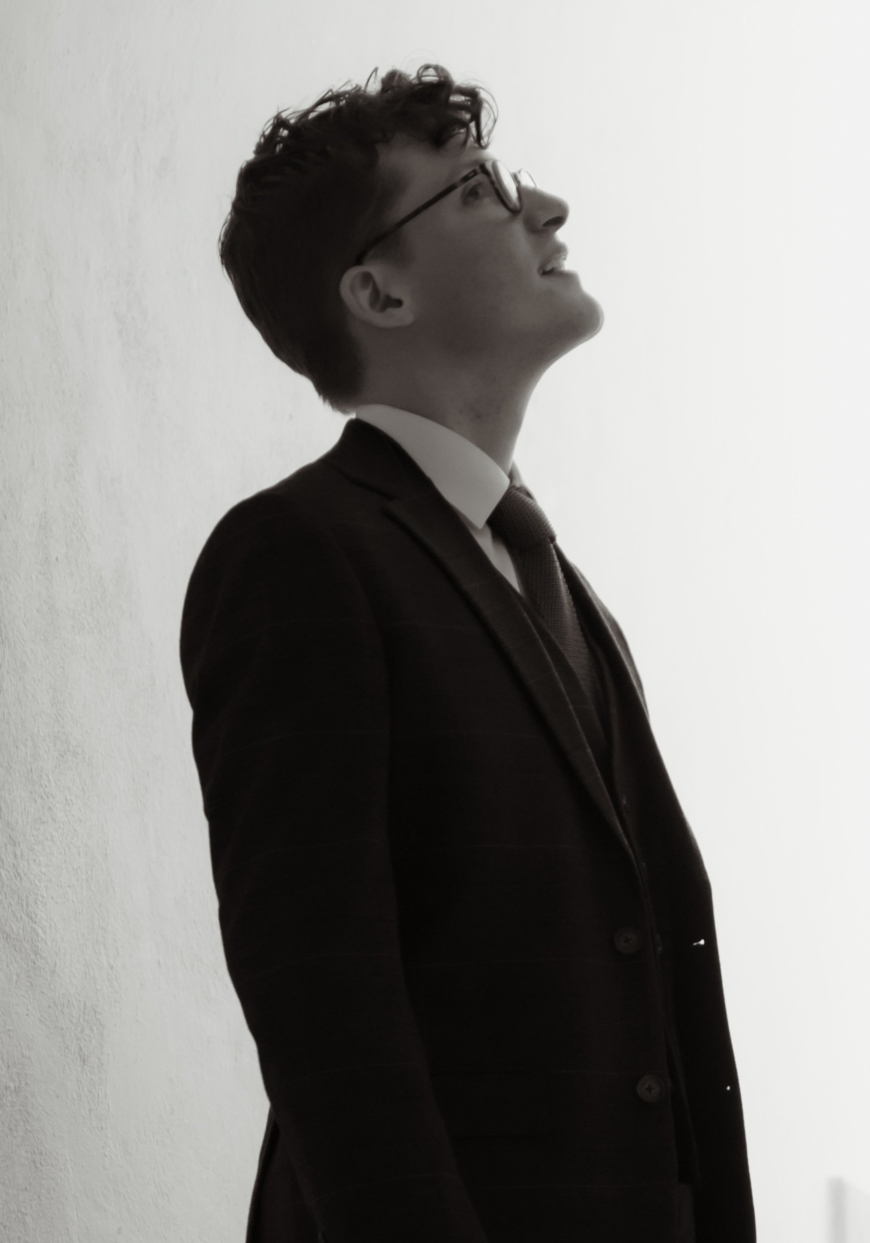 Profile of a young man with glasses in a business suit, smiling and looking upward, standing against a plain white wall.