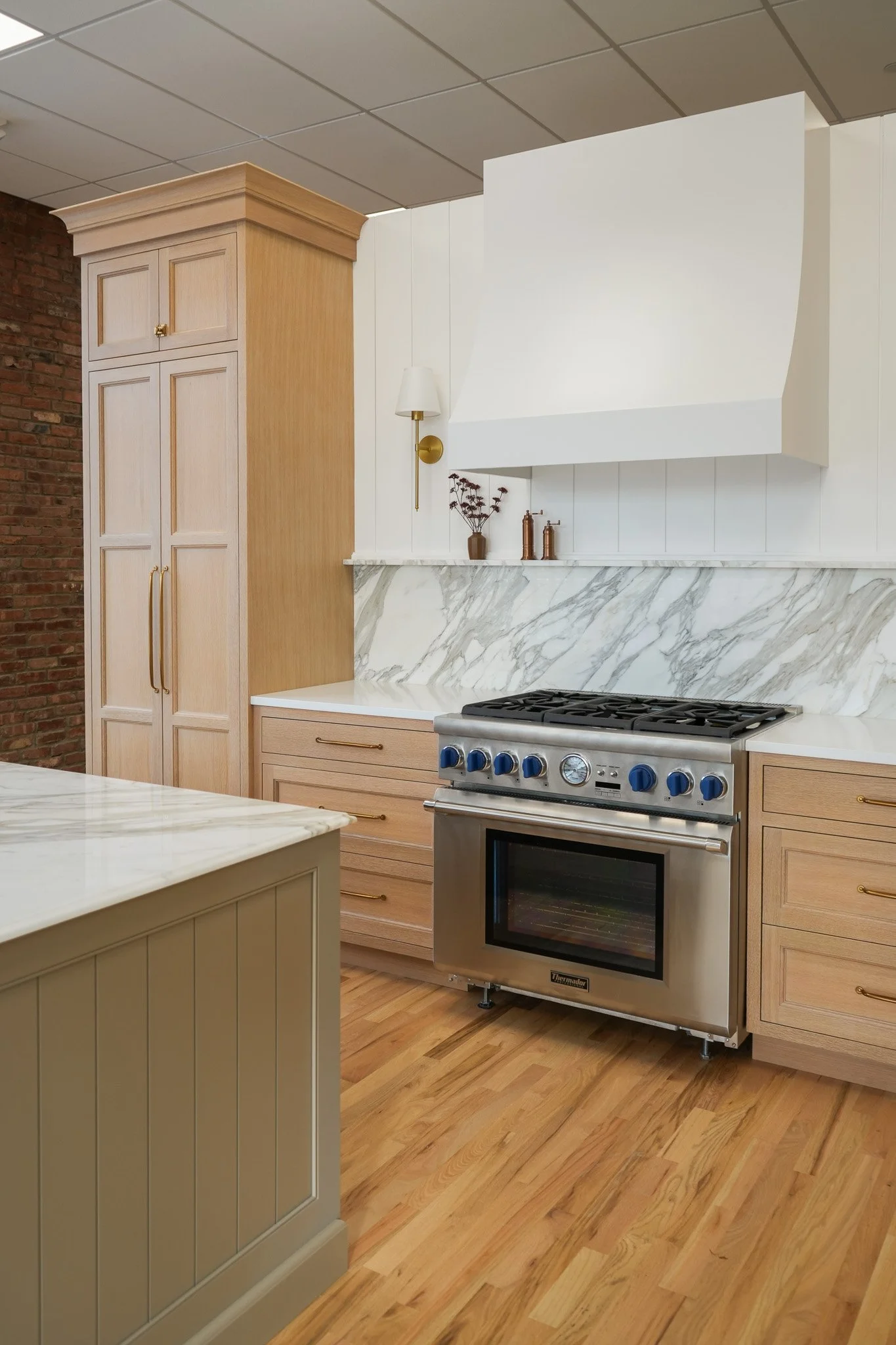 Modern kitchen with beige cabinetry, a marble countertop, a stainless steel oven, a white wall with a decorative vent, a wall-mounted lamp, and wooden flooring.