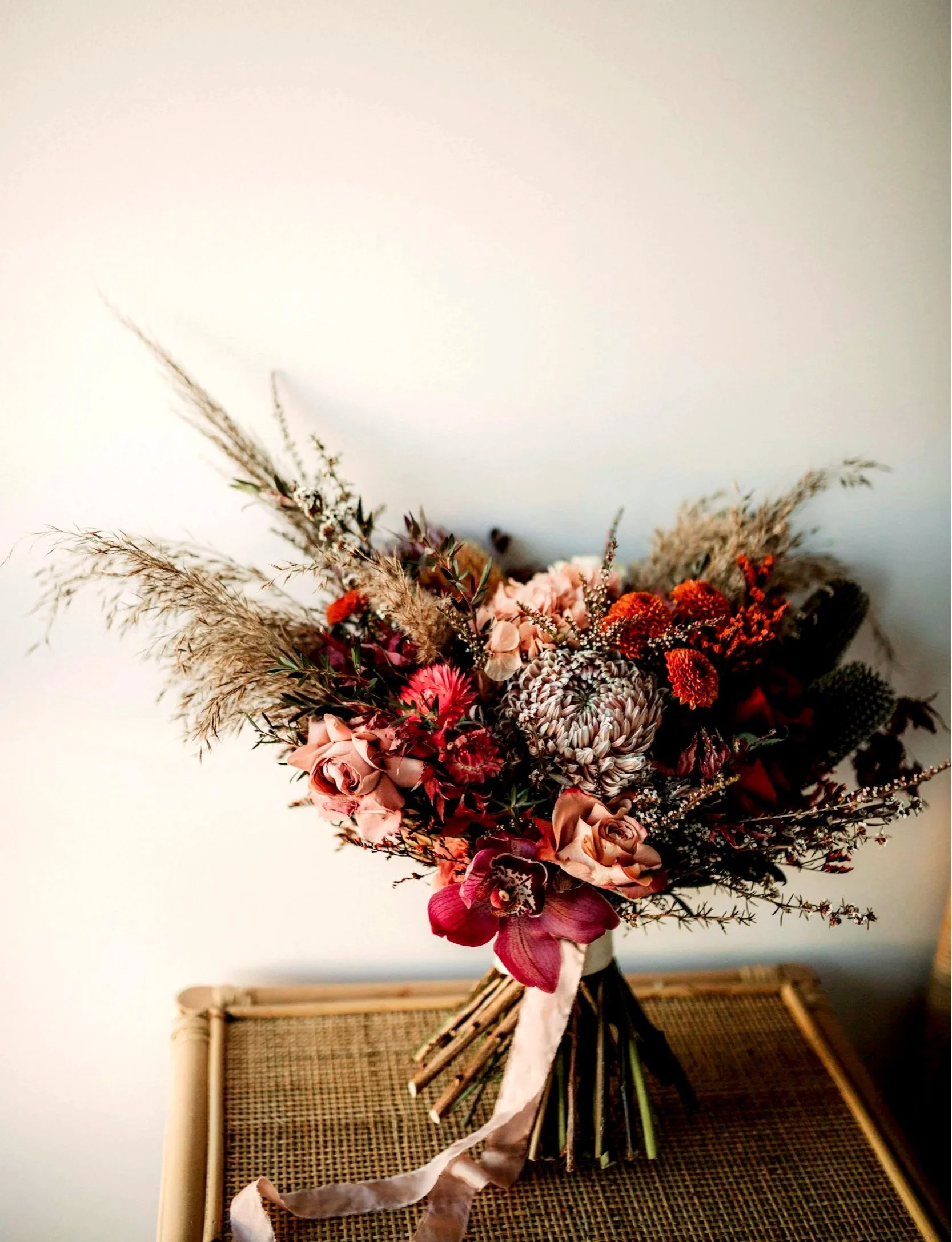 A bouquet of mixed flowers including roses, orchids, and dried grasses, wrapped with a pink ribbon, placed on a woven surface against a plain wall.