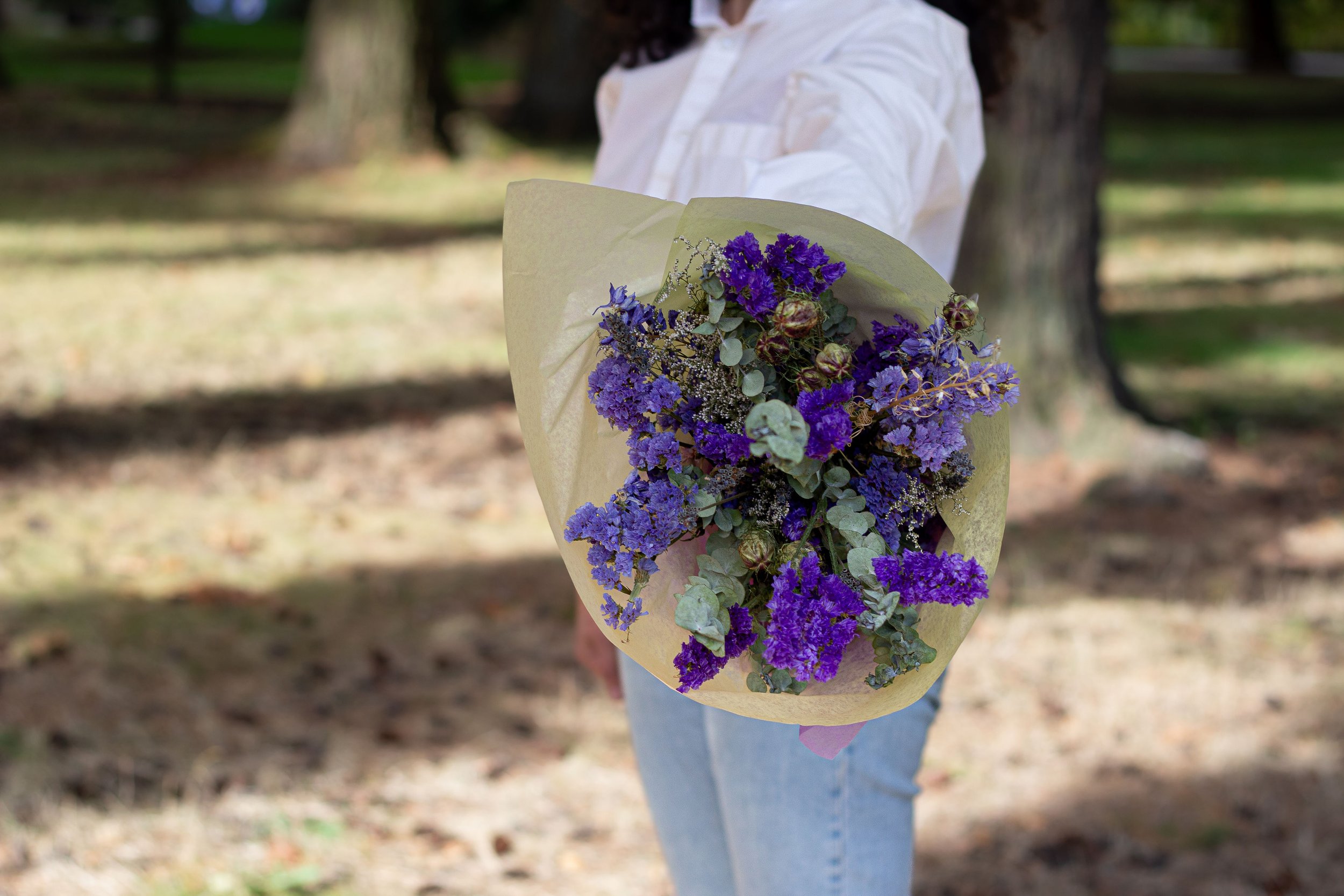 Person holding a bouquet of purple and blue flowers wrapped in light paper in a park with trees.