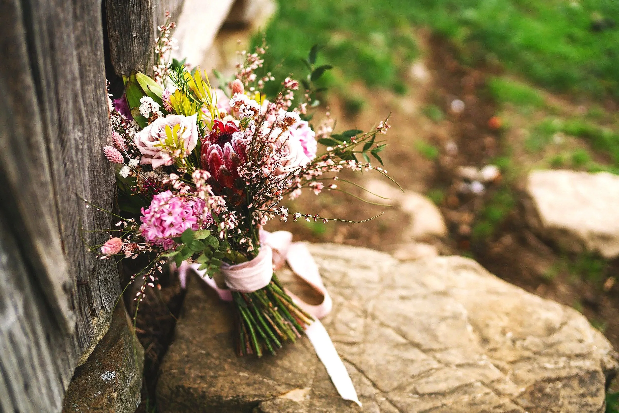 A bouquet of mixed flowers, including pink roses, purple carnations, yellow lilies, and other blooms, resting against a wooden fence on a stone bench outdoors.