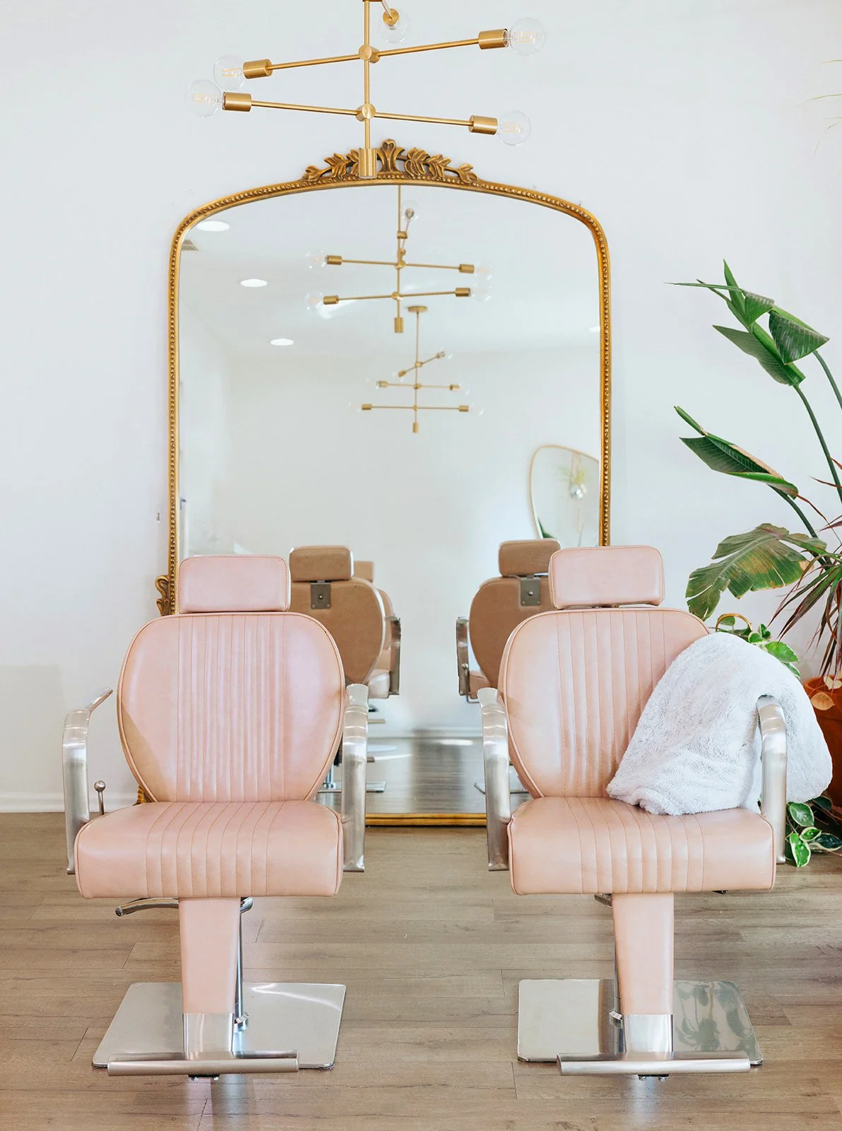 Vintage salon chairs with pink upholstery and metal armrests in front of a large ornate mirror, with a modern chandelier overhead and green plants on the side.