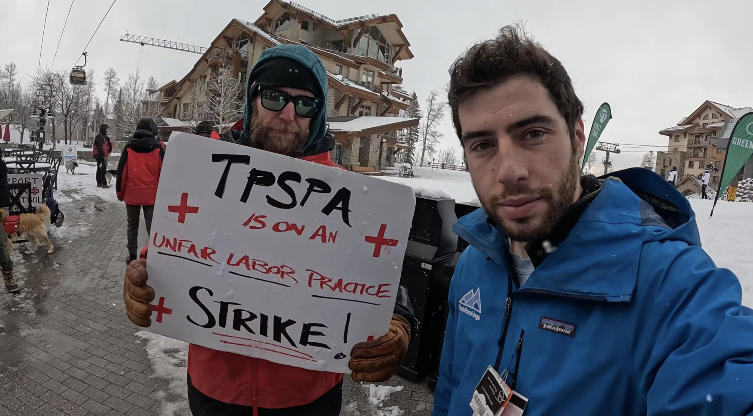 A reporter in a blue jacket standing with a striking ski patroller holding a picketing sign, on a snowy day in a resort village in Telluride, Colorado