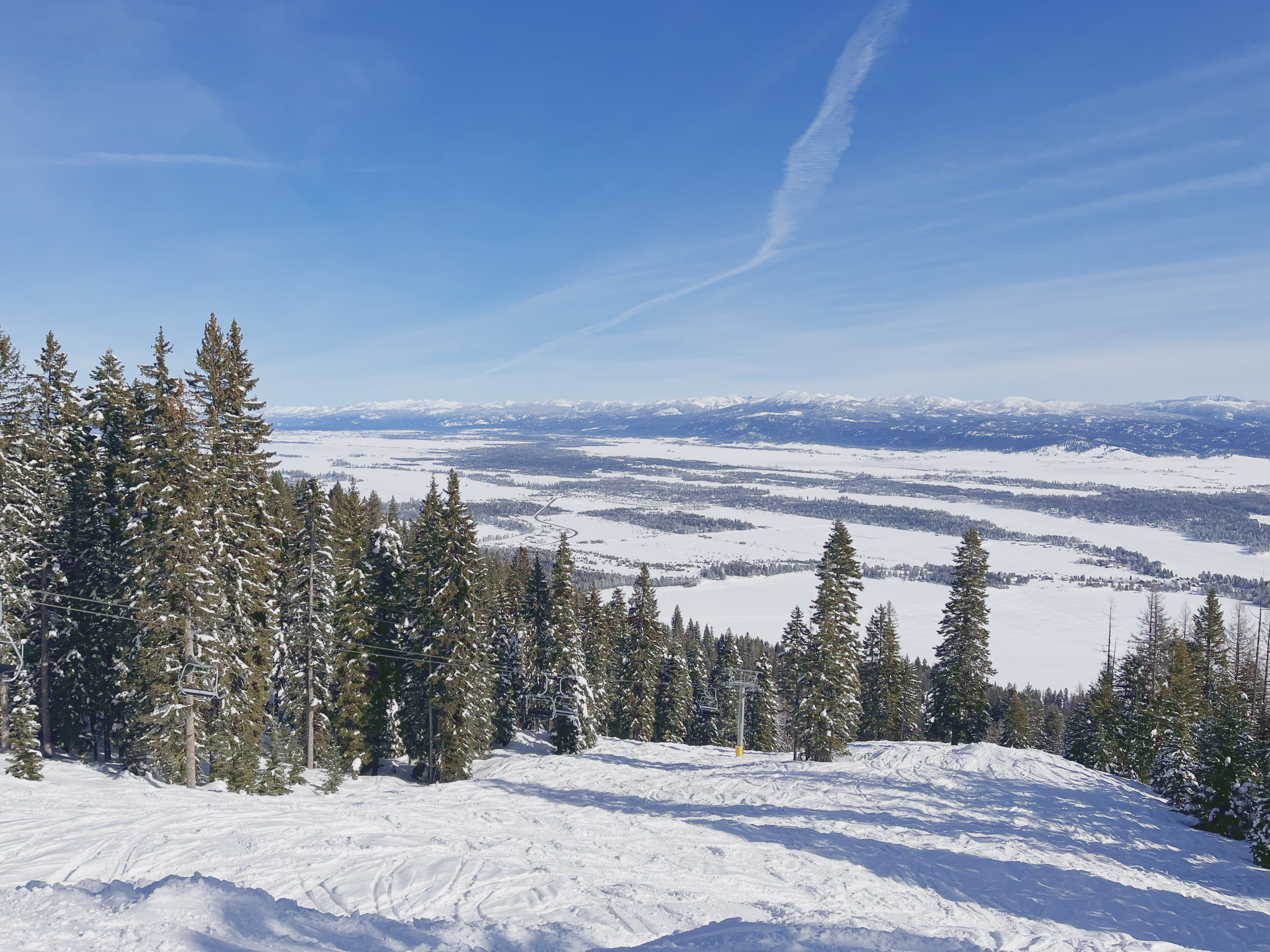 Panoramic view from a snowy ski slope of trees, a chairlift, and a snowy plain and mountain range in the distance on a clear sunny day at Tamarack ski resort, Idaho