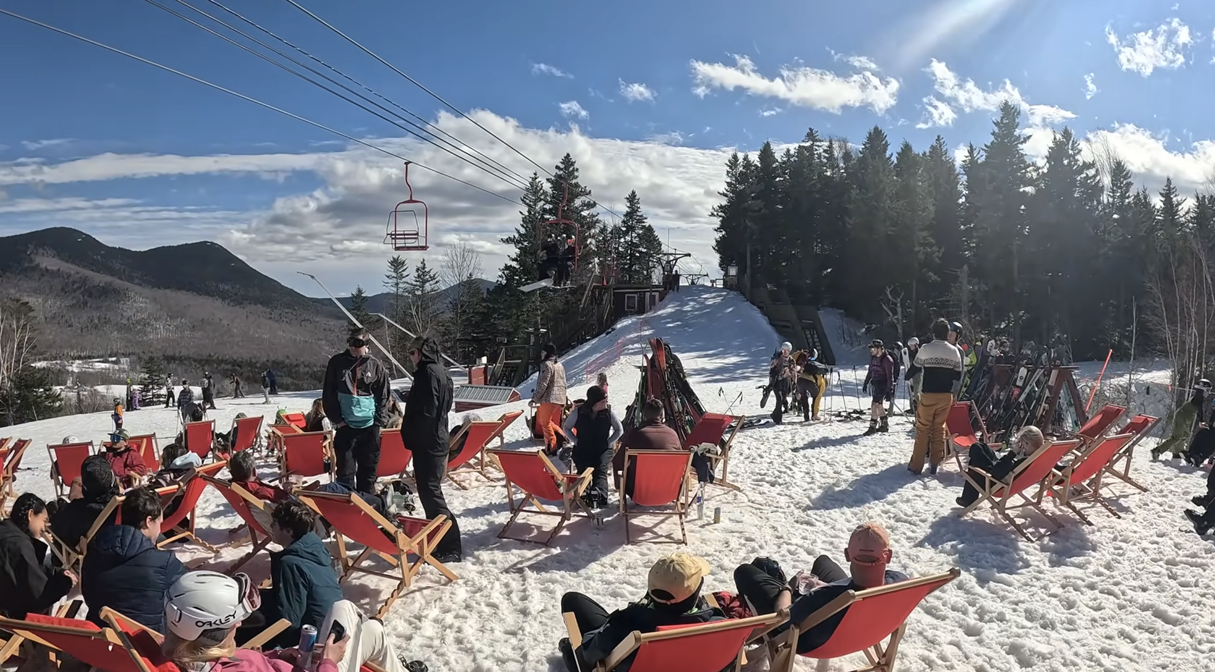 People sit in lounge chairs on snow with an orange chairlift passing over their heads on a sunny day at Black Mountain, NH