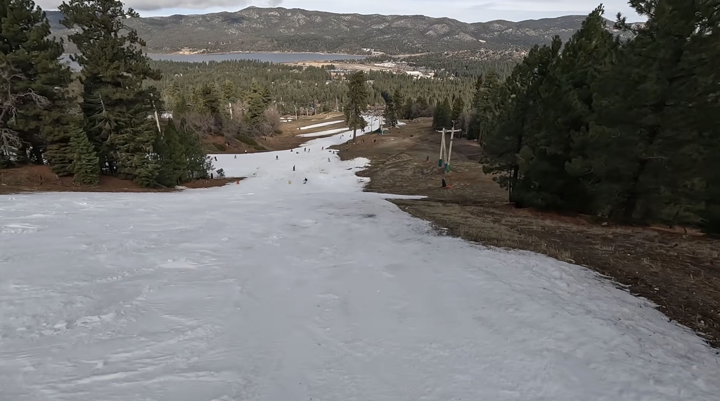Skiing down a narrow slope with dirt on both sides and a lake and forest in the background at Big Bear, CA
