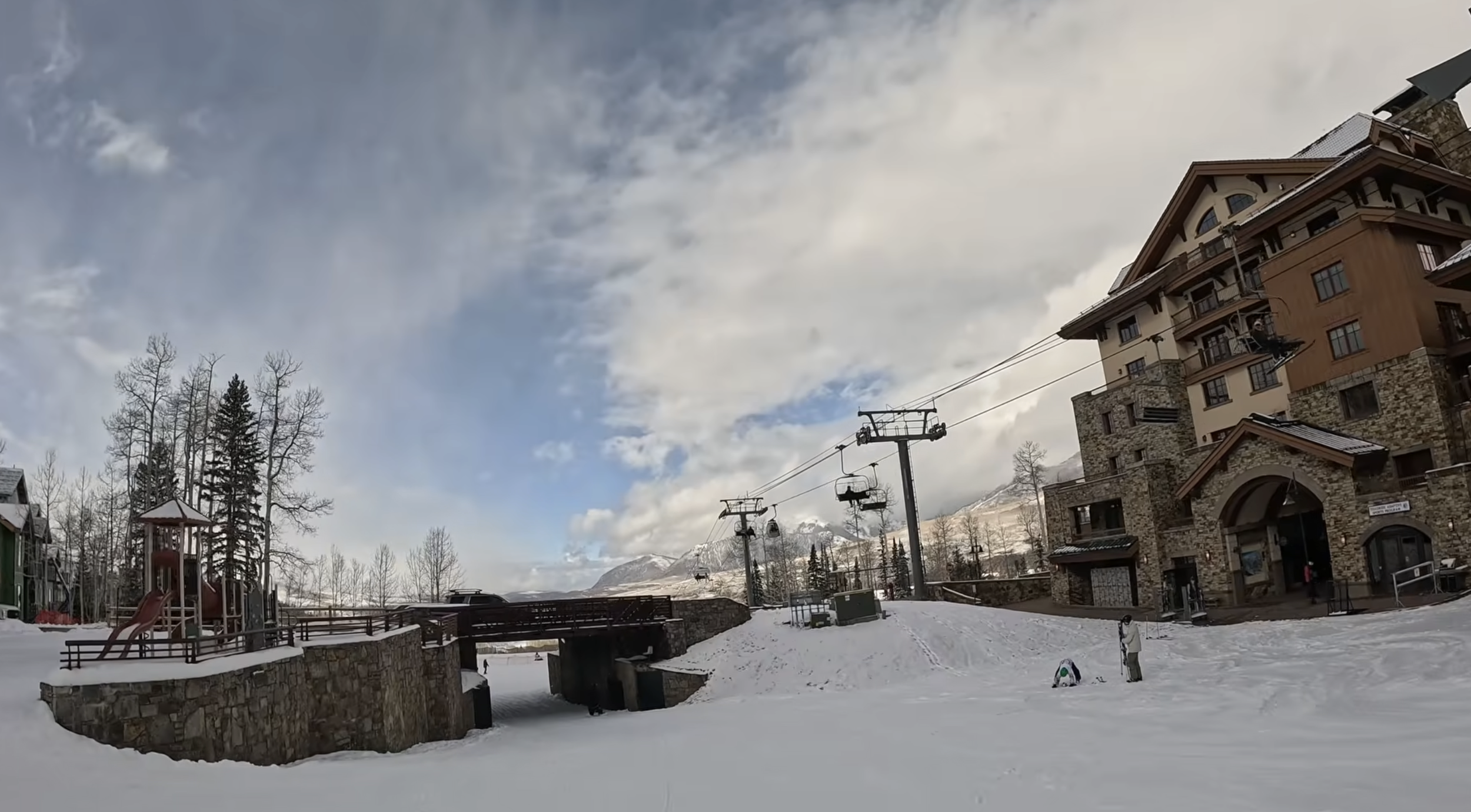 A ski lift running past a resort condo building, which is connected to a playground on the other side by a bridge. A gentle ski slope runs underneath the bridge
