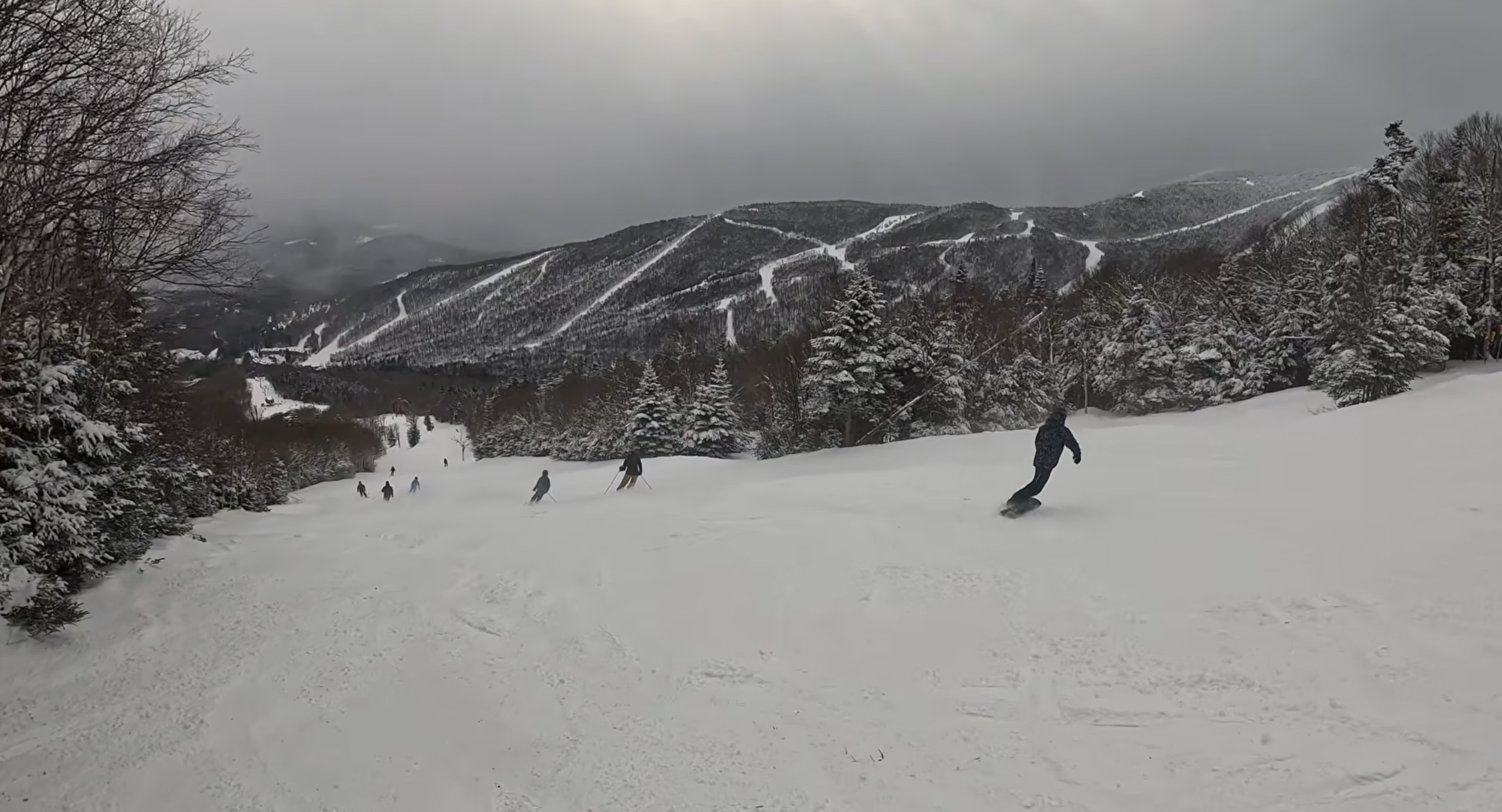 Skiing down a steady ski slope with other skiers and snowboarders and a view of many ski runs on a mountain in the distance on a cloudy day at Sugarbush ski resort, VT