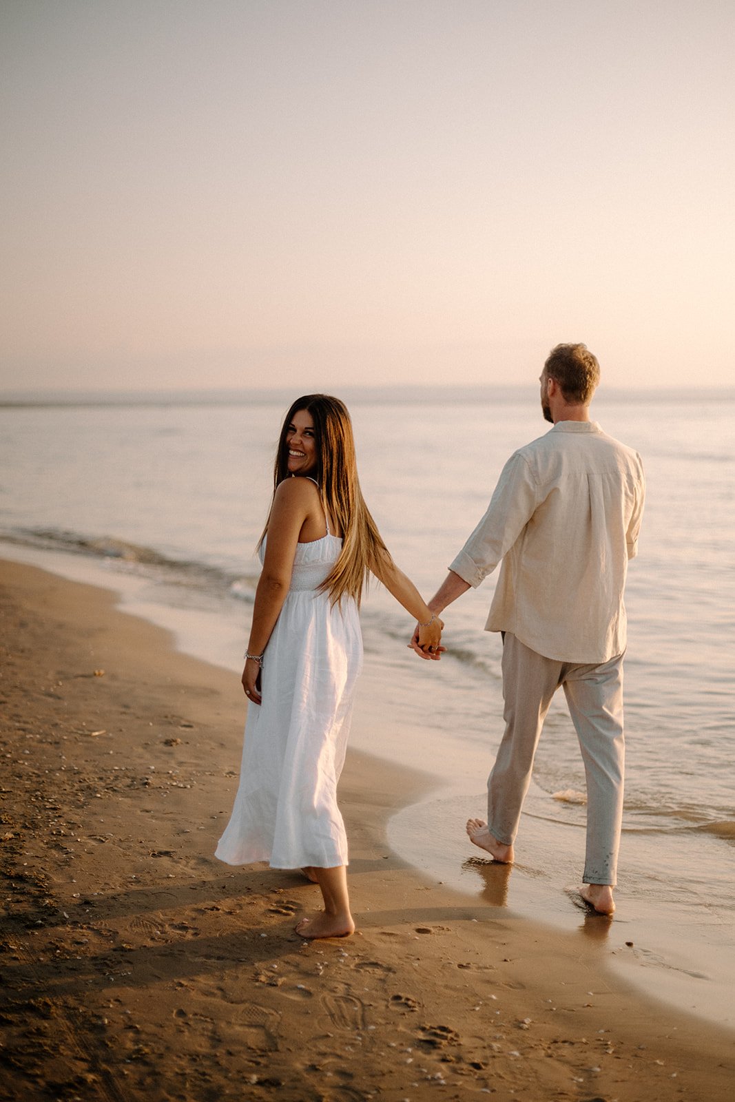 A couple holding hands and walking barefoot on the beach at sunset, with the woman smiling at the camera.